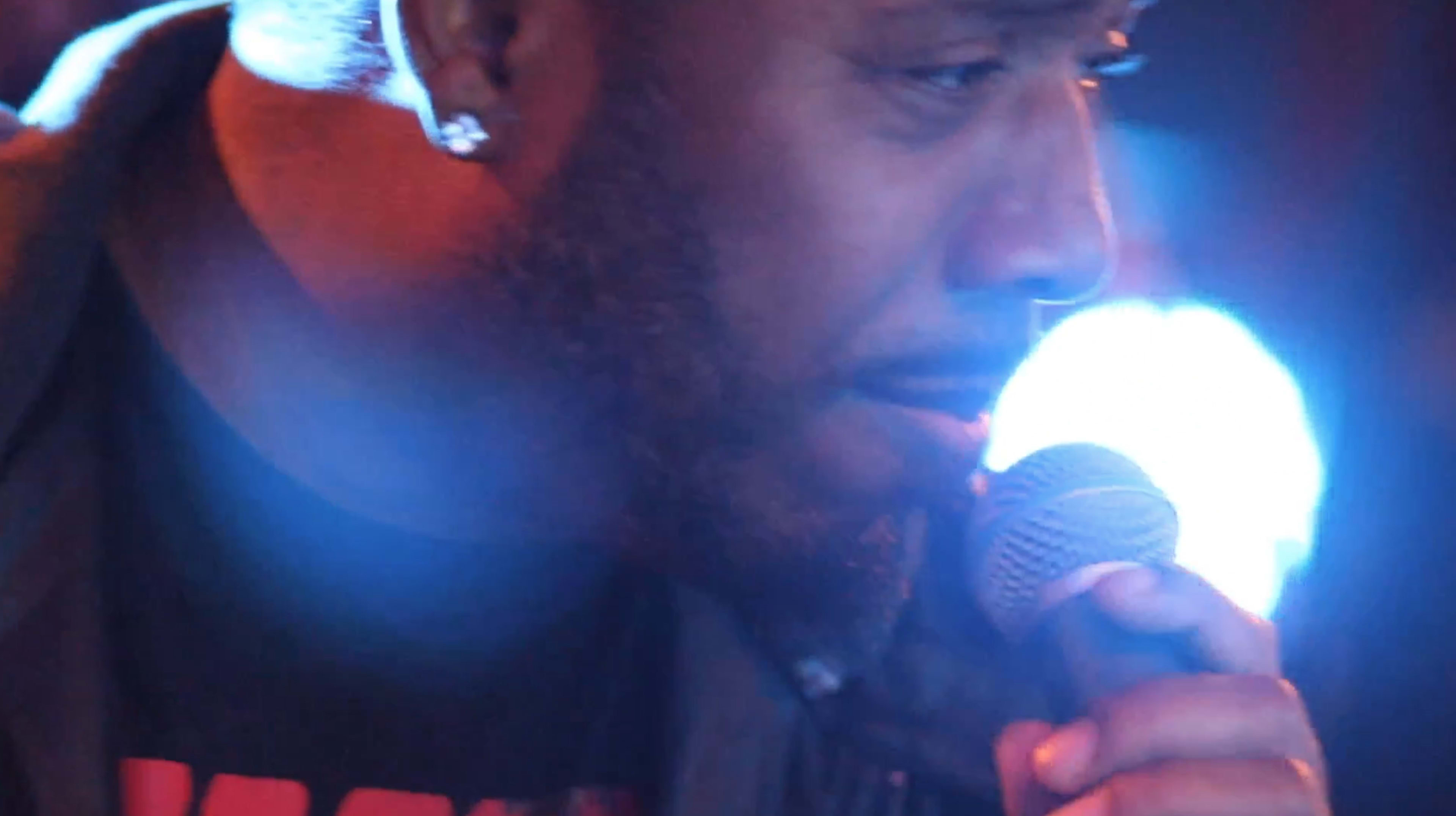 A performer holds a microphone close to his face while illuminated by vibrant stage lights.