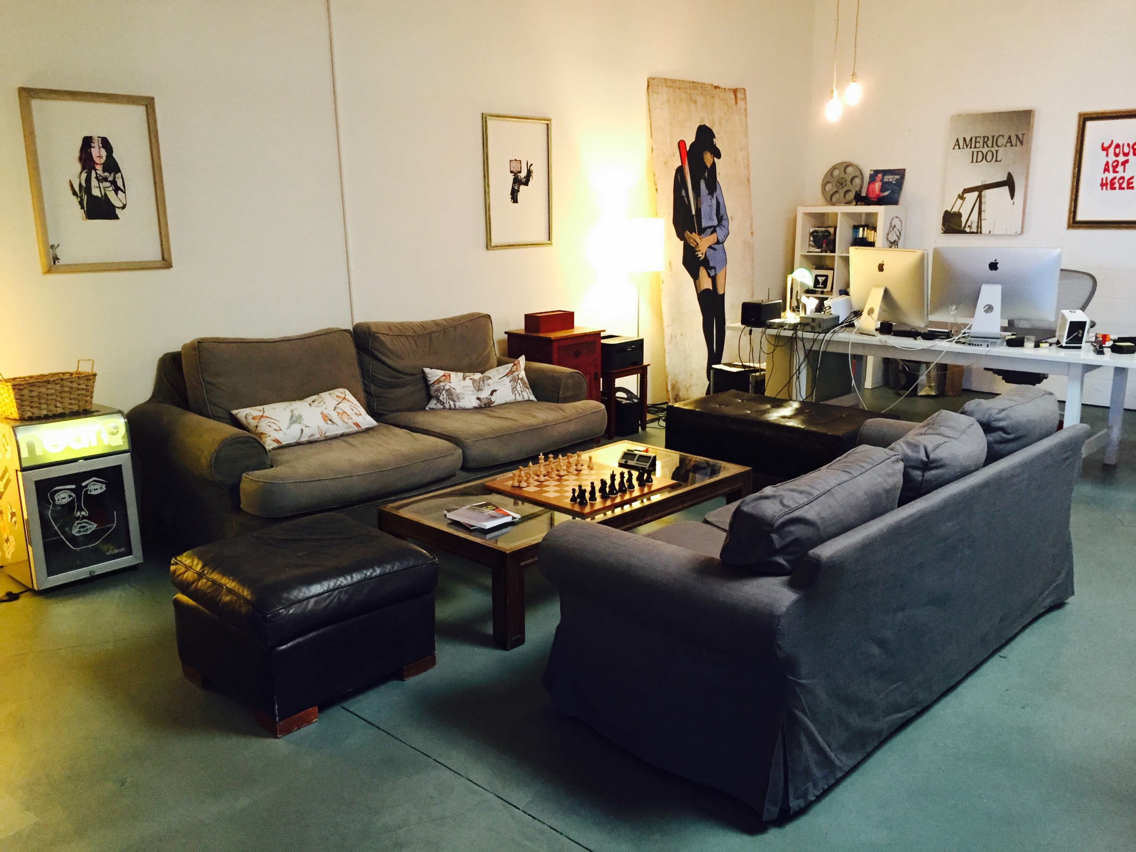 The image shows a cozy living room with two sofas, a coffee table featuring a chess set, and a desk area with a computer setup against a plain wall.