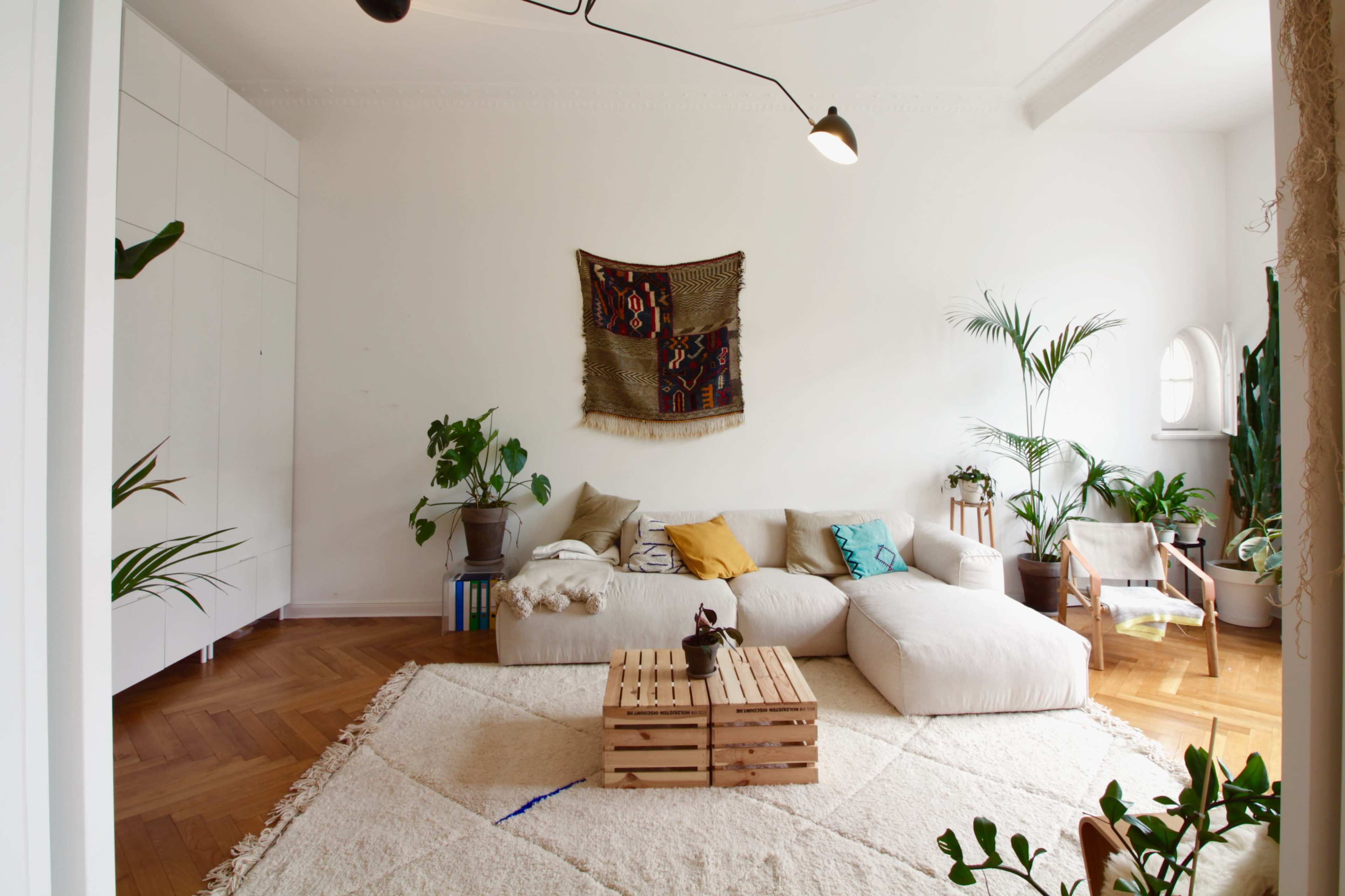 The image shows a bright living room with a light-colored sectional sofa, a wooden coffee table made of crates, and various indoor plants surrounding the space.