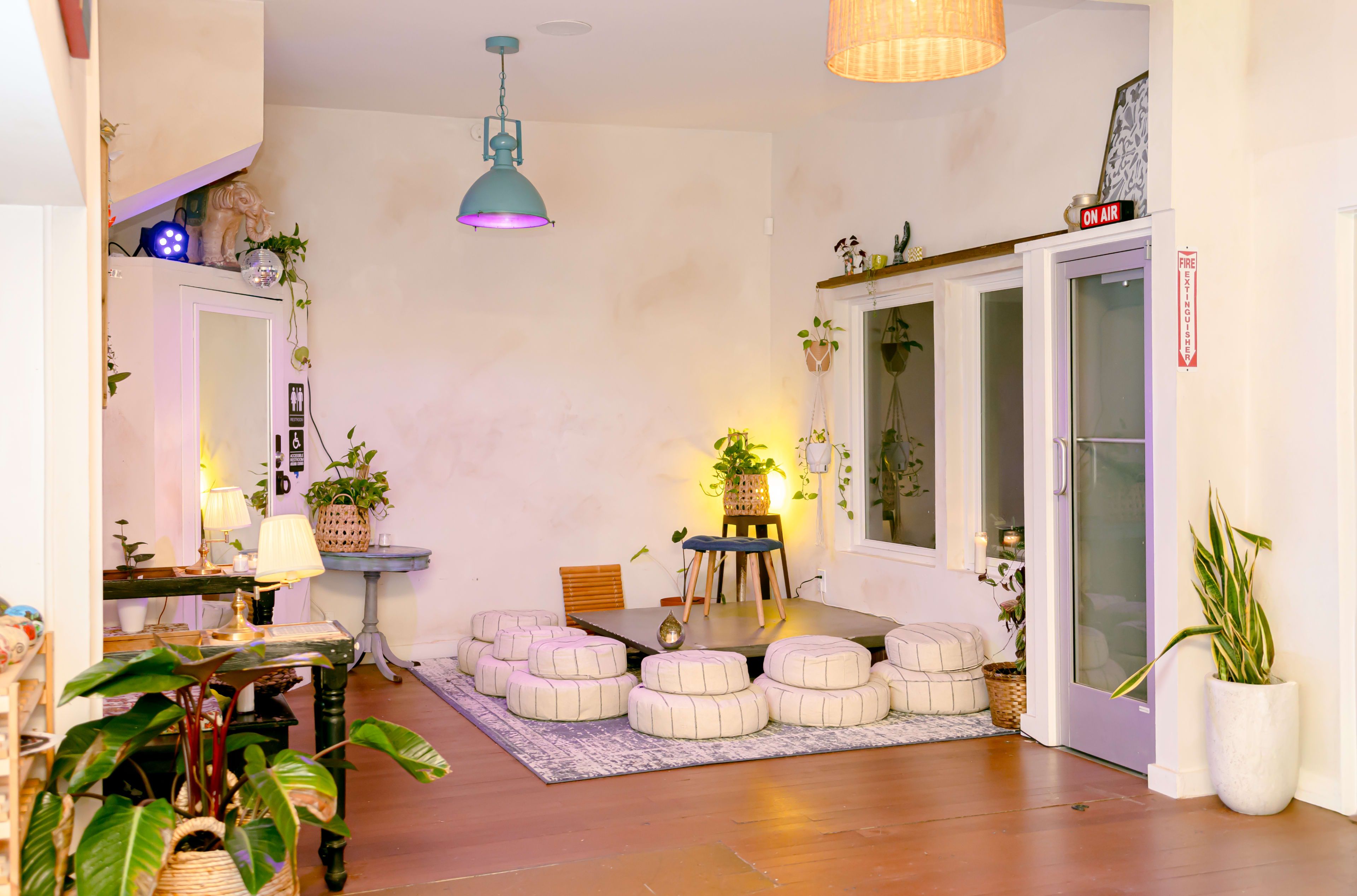 The image shows a cozy room decorated with plants, a circular seating area of cushioned ottomans, and a bright pendant light hanging from the ceiling.