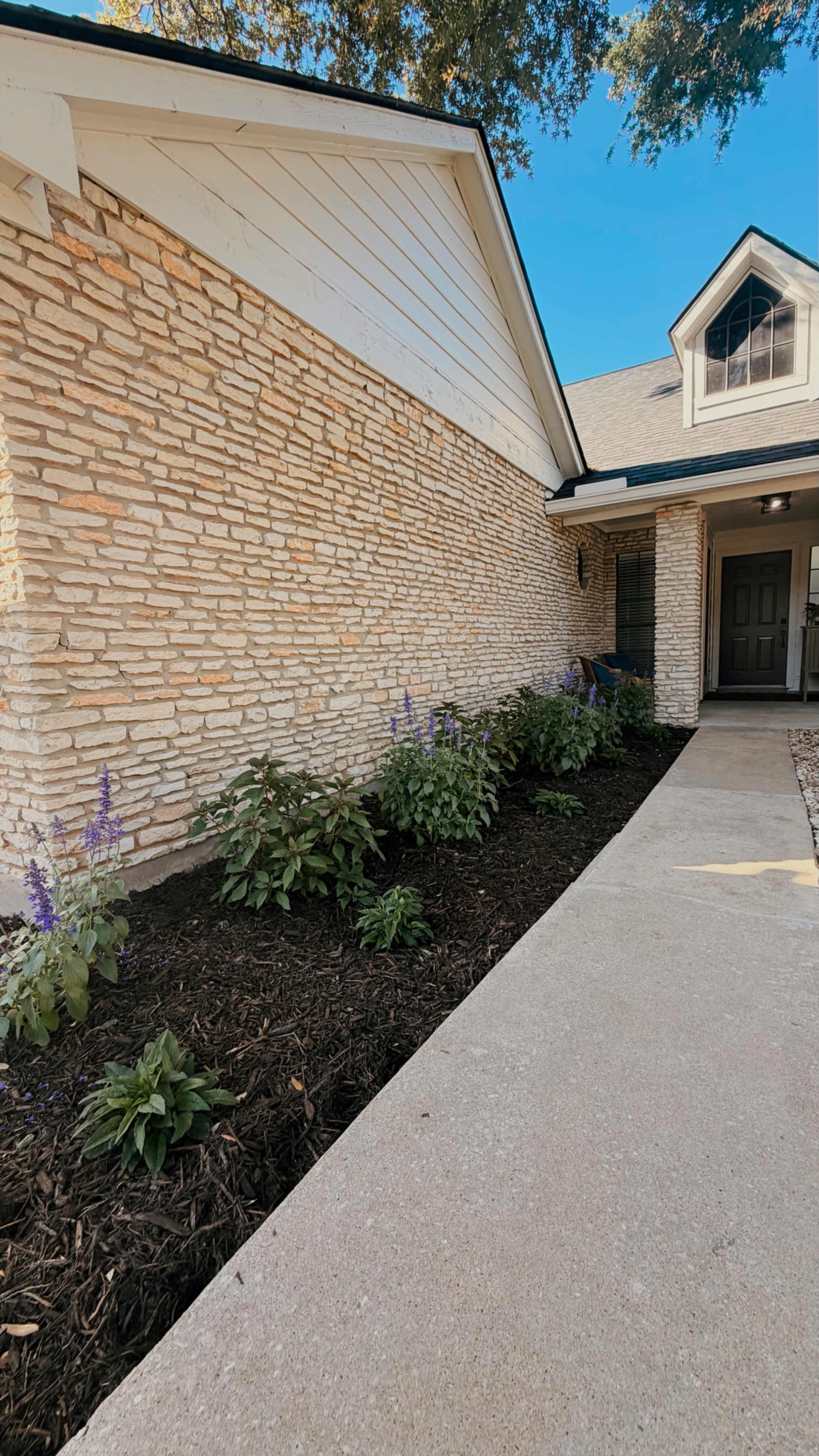 The image shows a stone and siding exterior of a house with a garden bed featuring purple flowers and dark mulch along a concrete walkway.