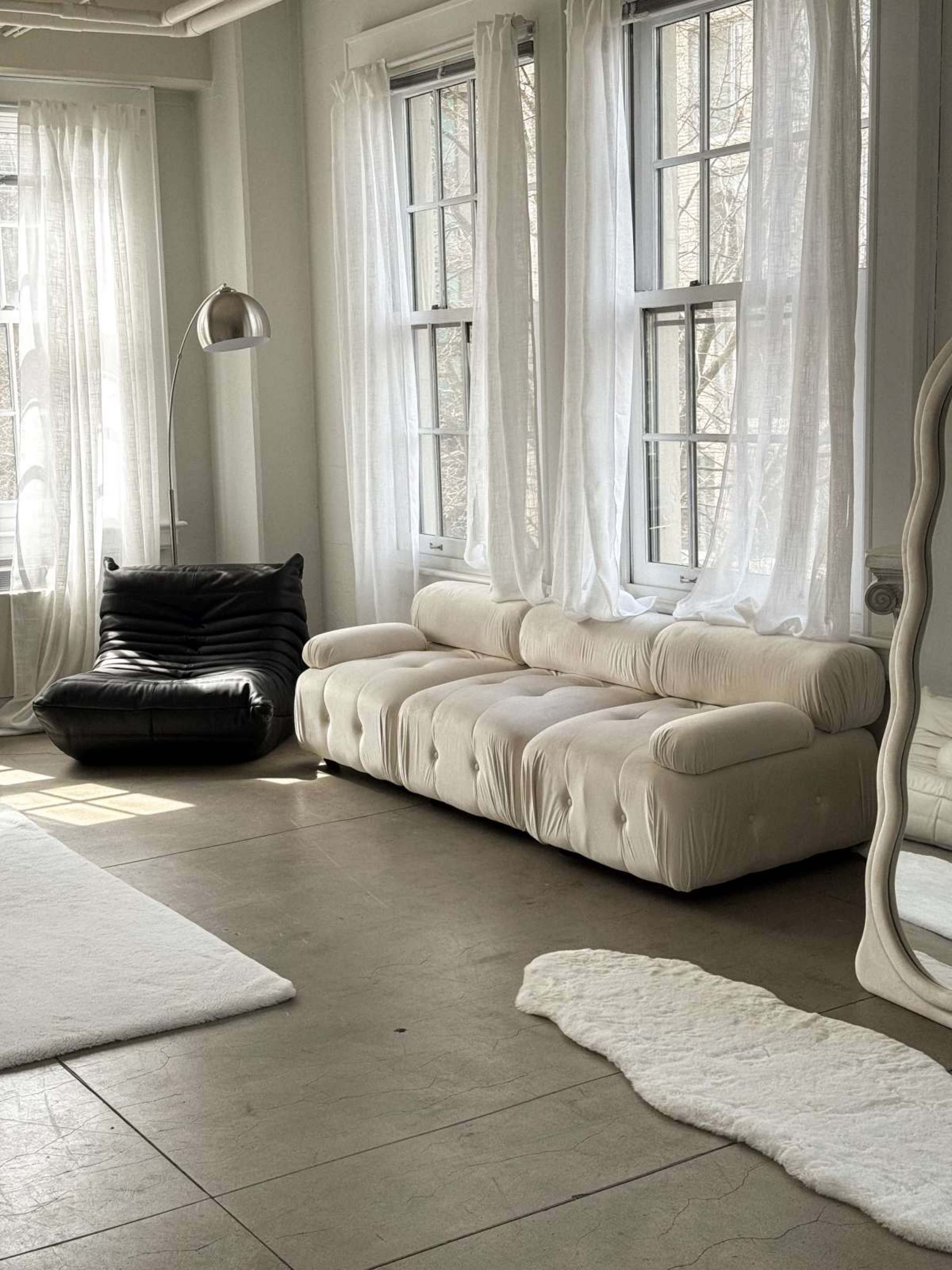 The image shows a modern, minimalist living space featuring a white, textured sofa, a black lounge chair, and large windows with sheer white curtains.