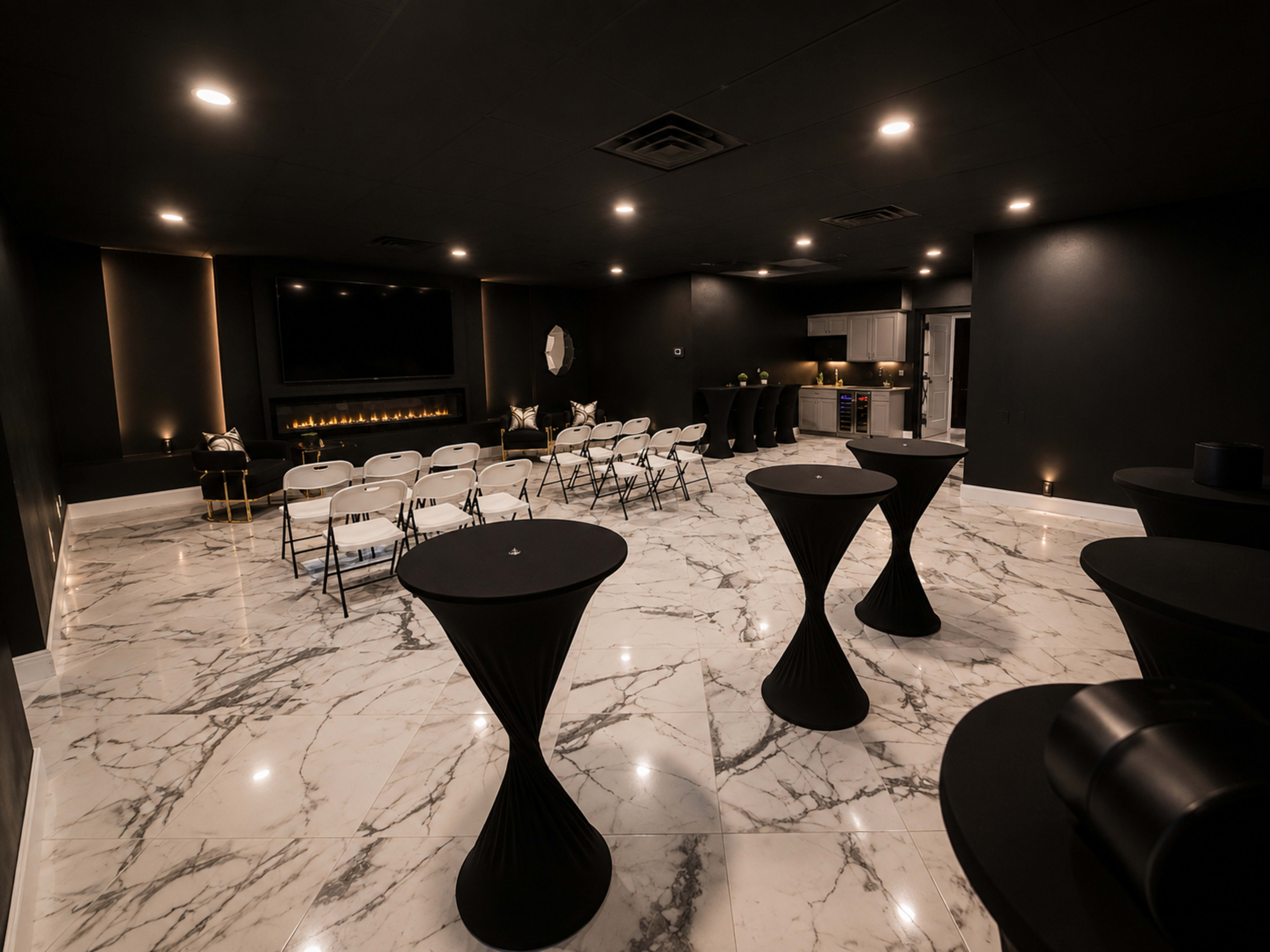 The image shows a modern event space with a marble floor, black walls, and rows of folding chairs facing a large screen, accompanied by cocktail tables scattered throughout.