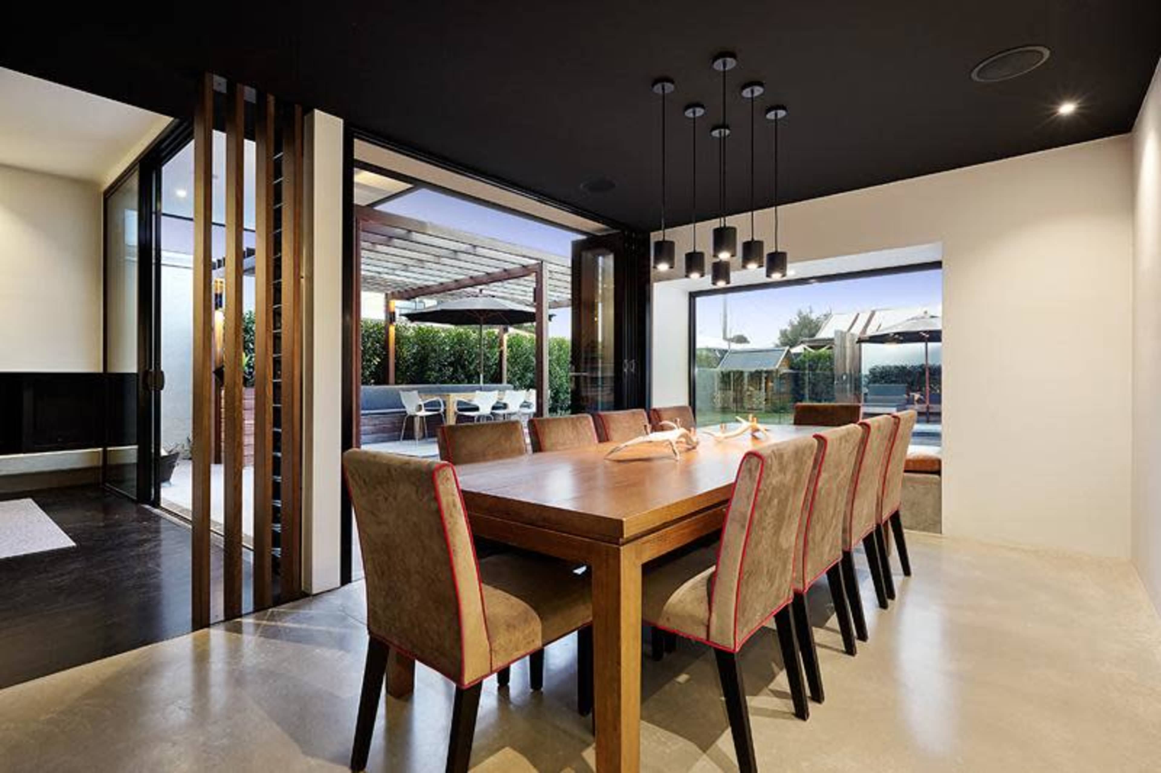 A dining area features a long wooden table surrounded by upholstered chairs, with large glass doors opening to an outdoor patio.