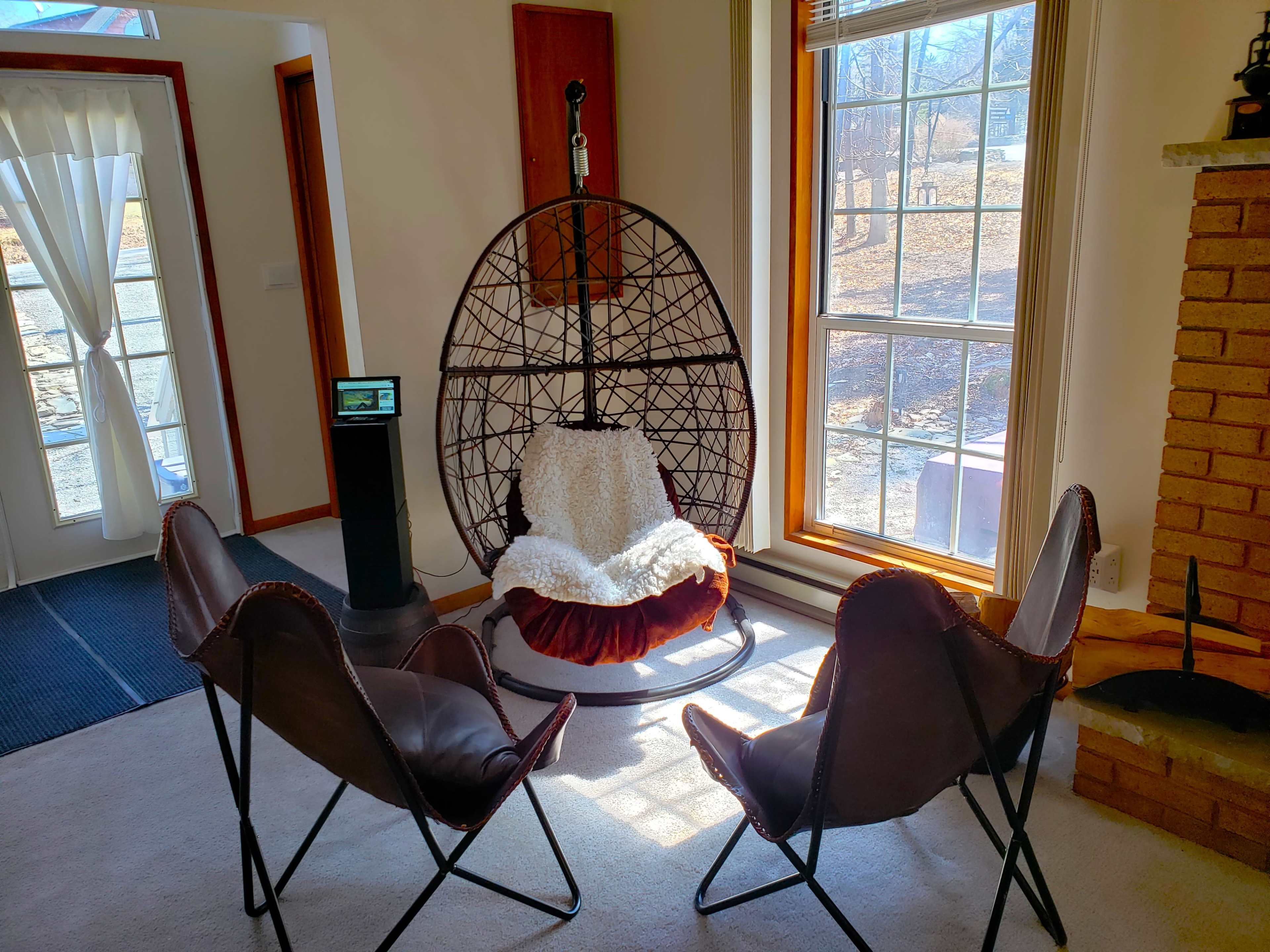 The image shows a cozy room featuring a large hanging chair surrounded by three leather chairs, with natural light streaming through a nearby window.