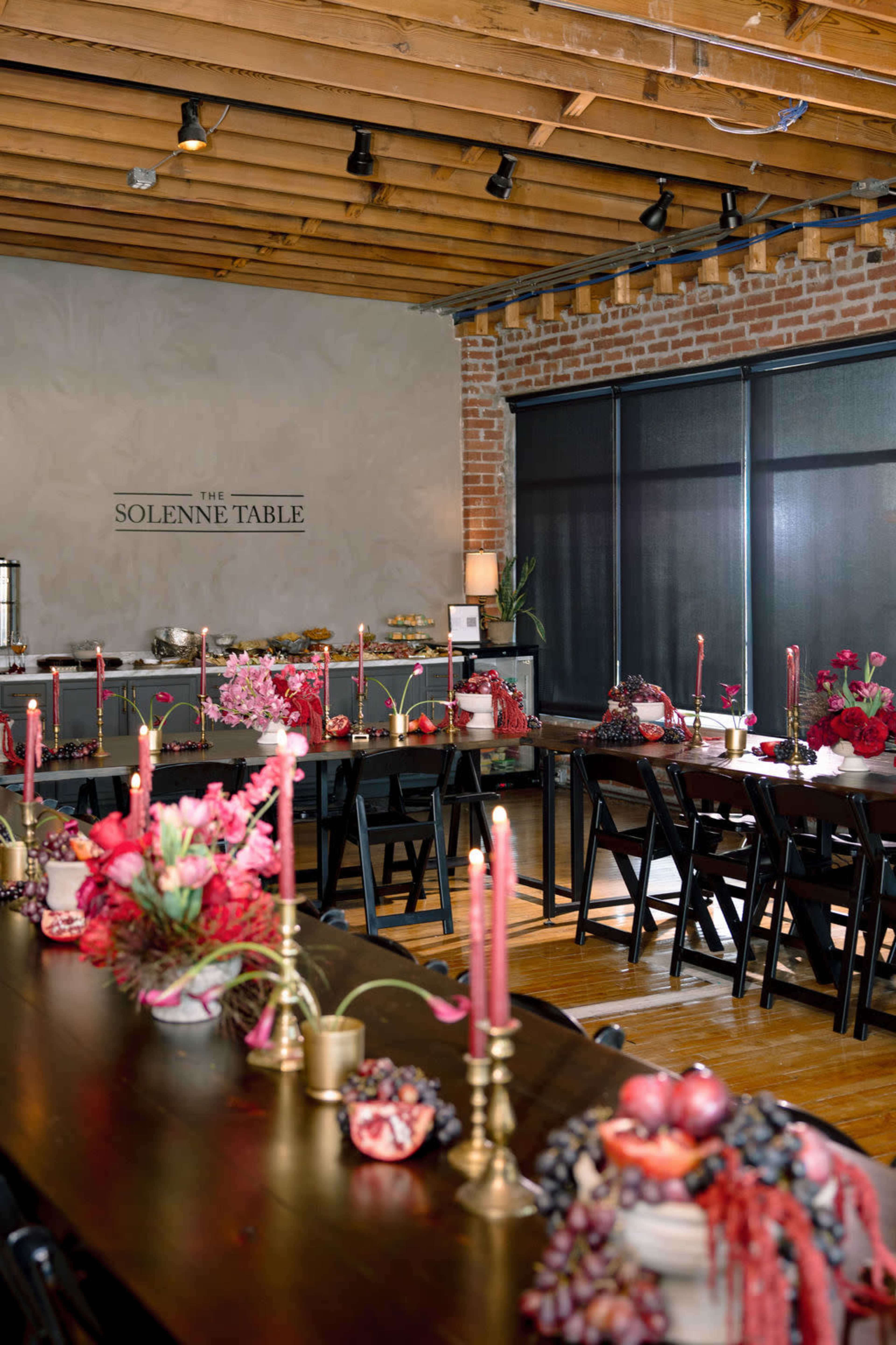 The image depicts a dining area with long tables adorned with candles, floral arrangements, and decorative items, set against a backdrop of exposed brick walls and wooden beams.