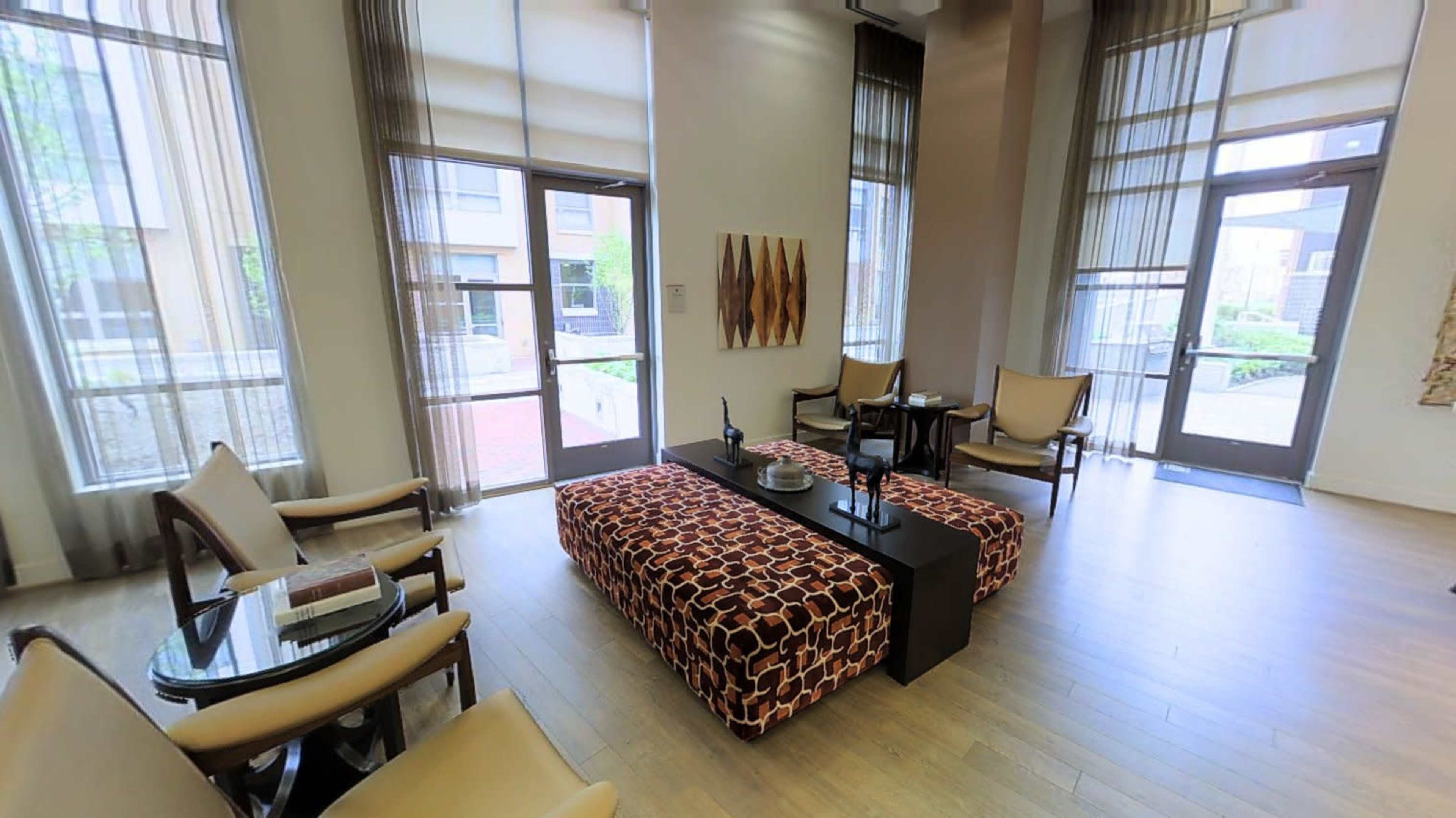 A modern living space with large windows, light-colored wood flooring, and minimalist furniture, including a patterned coffee table and armchairs.