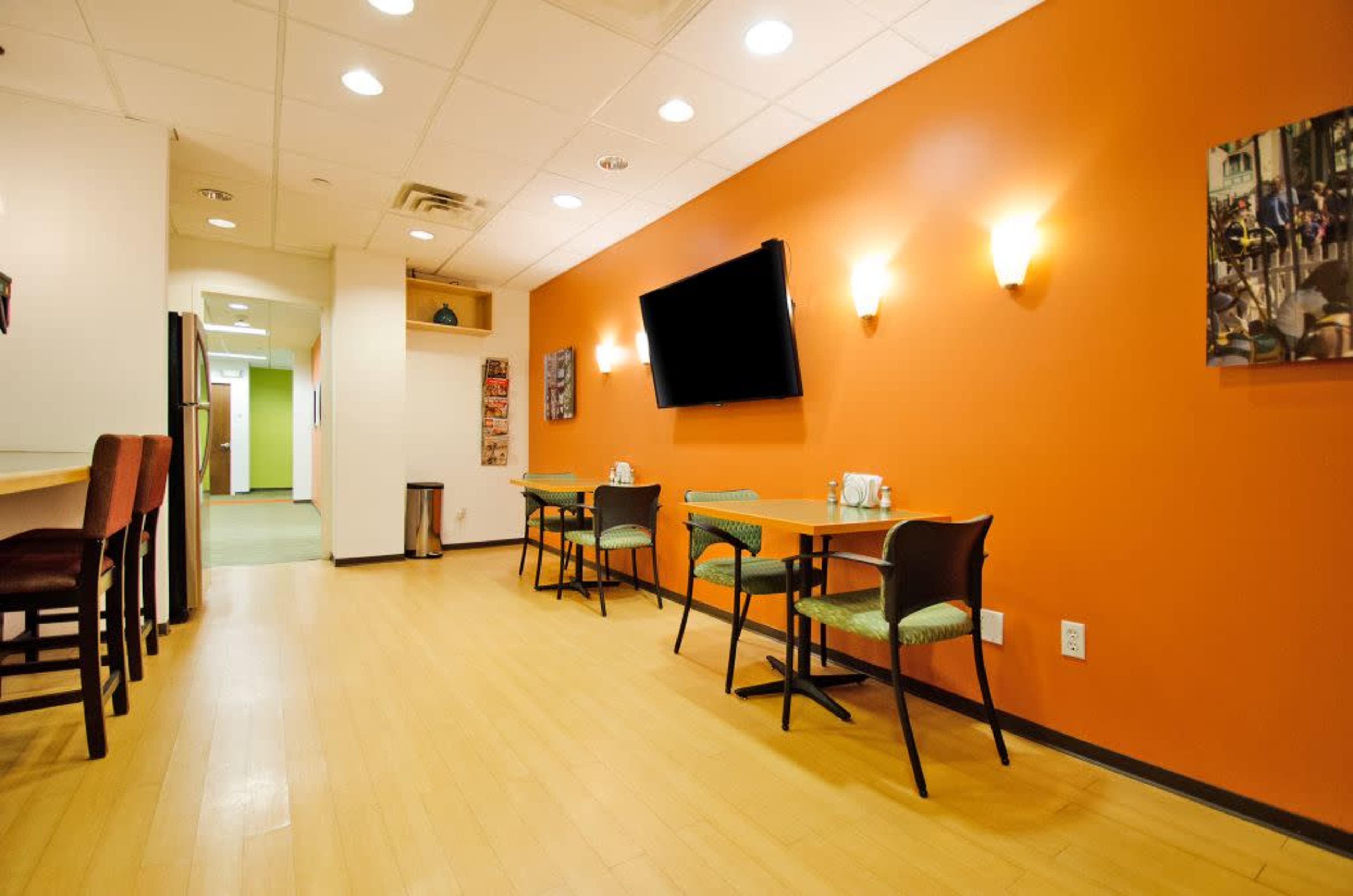 A room with orange walls, hardwood flooring, two tables with chairs, and a wall-mounted television.