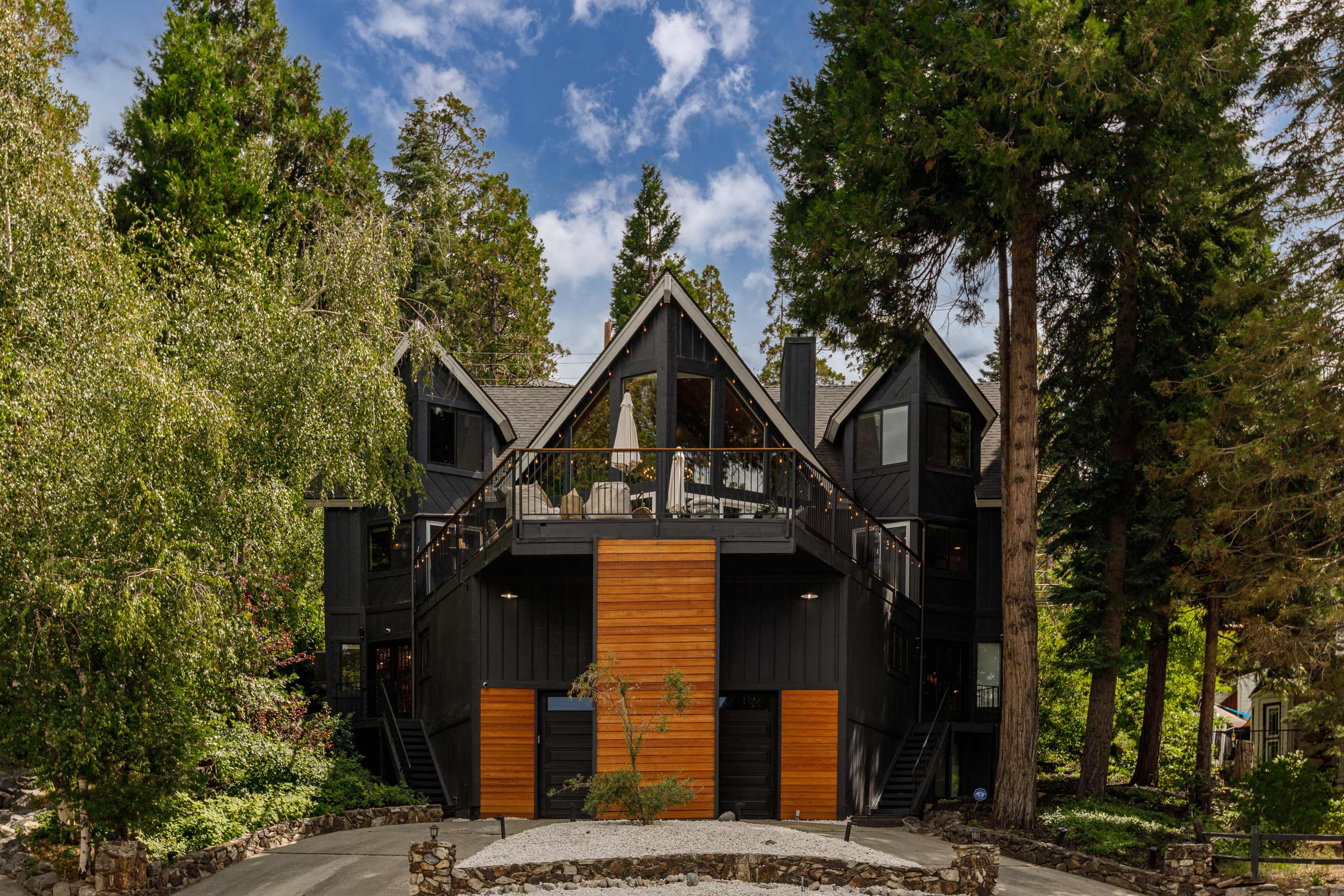 A modern black chalet-style house with wooden accents is surrounded by tall trees and greenery.
