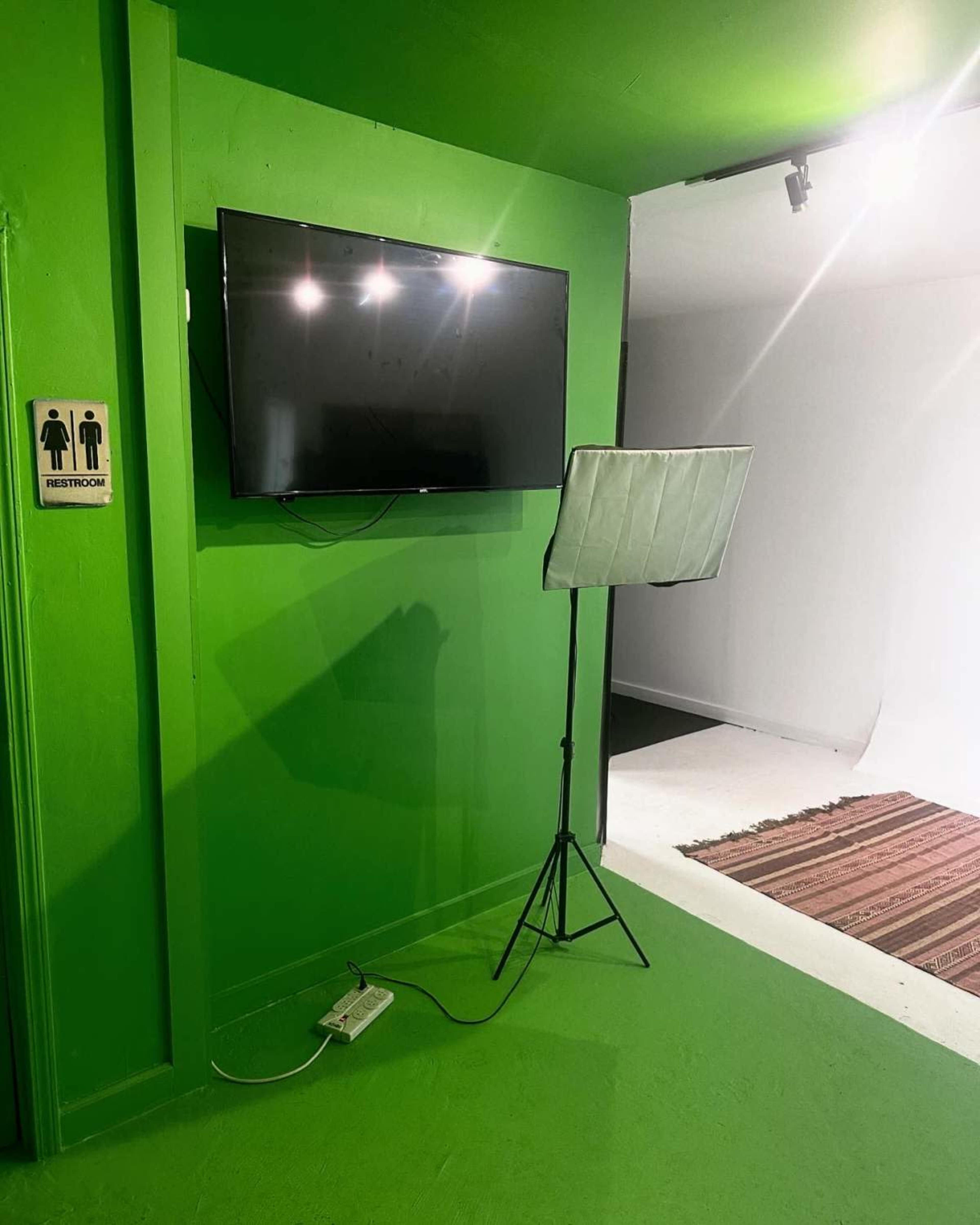 A green-walled room features a mounted television and a folded screen on a stand, with a restroom sign nearby.