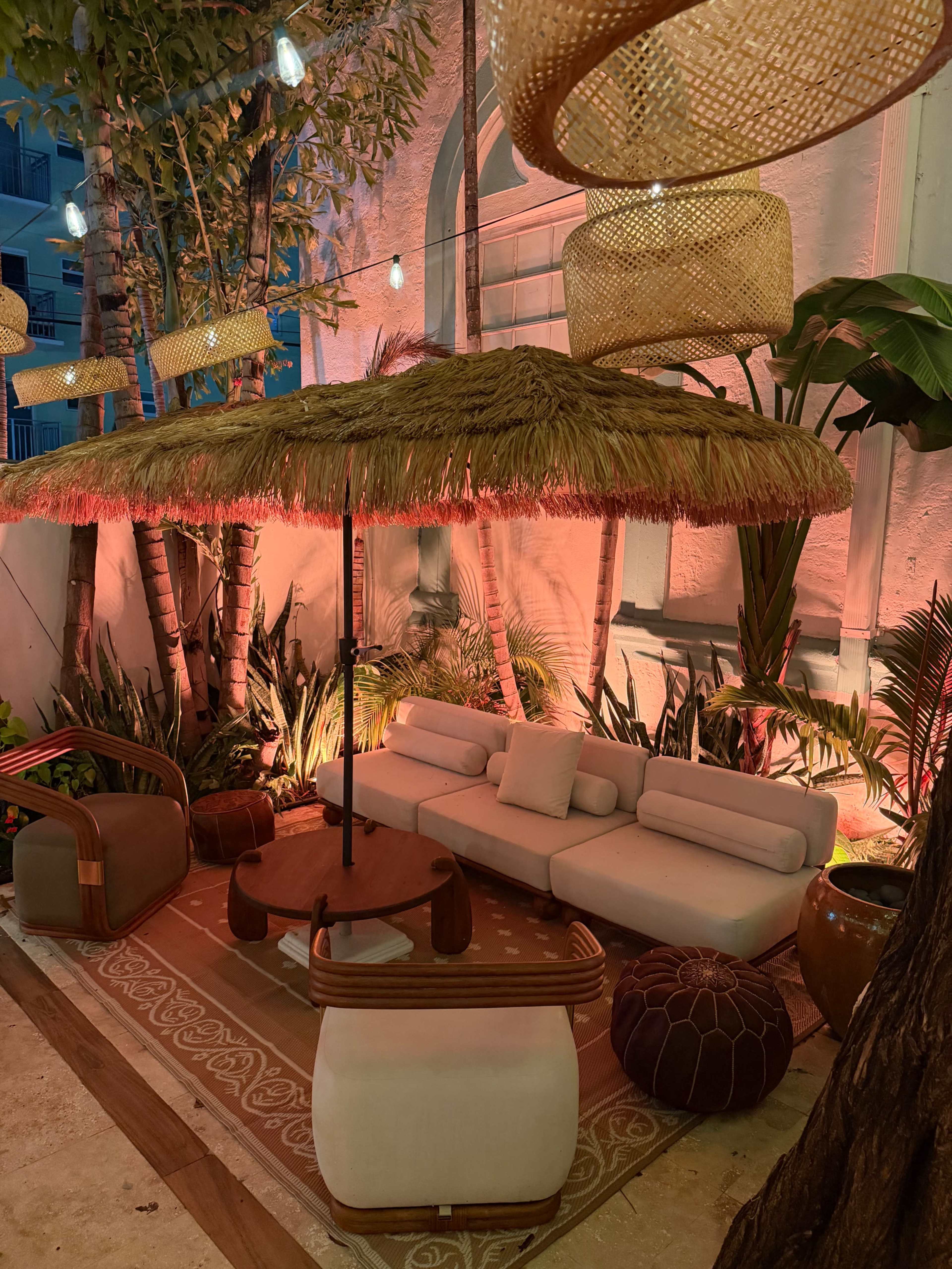 The image shows a cozy outdoor lounge area with a thatched umbrella, a light-colored sectional sofa, and decorative plants, all illuminated with warm lighting.