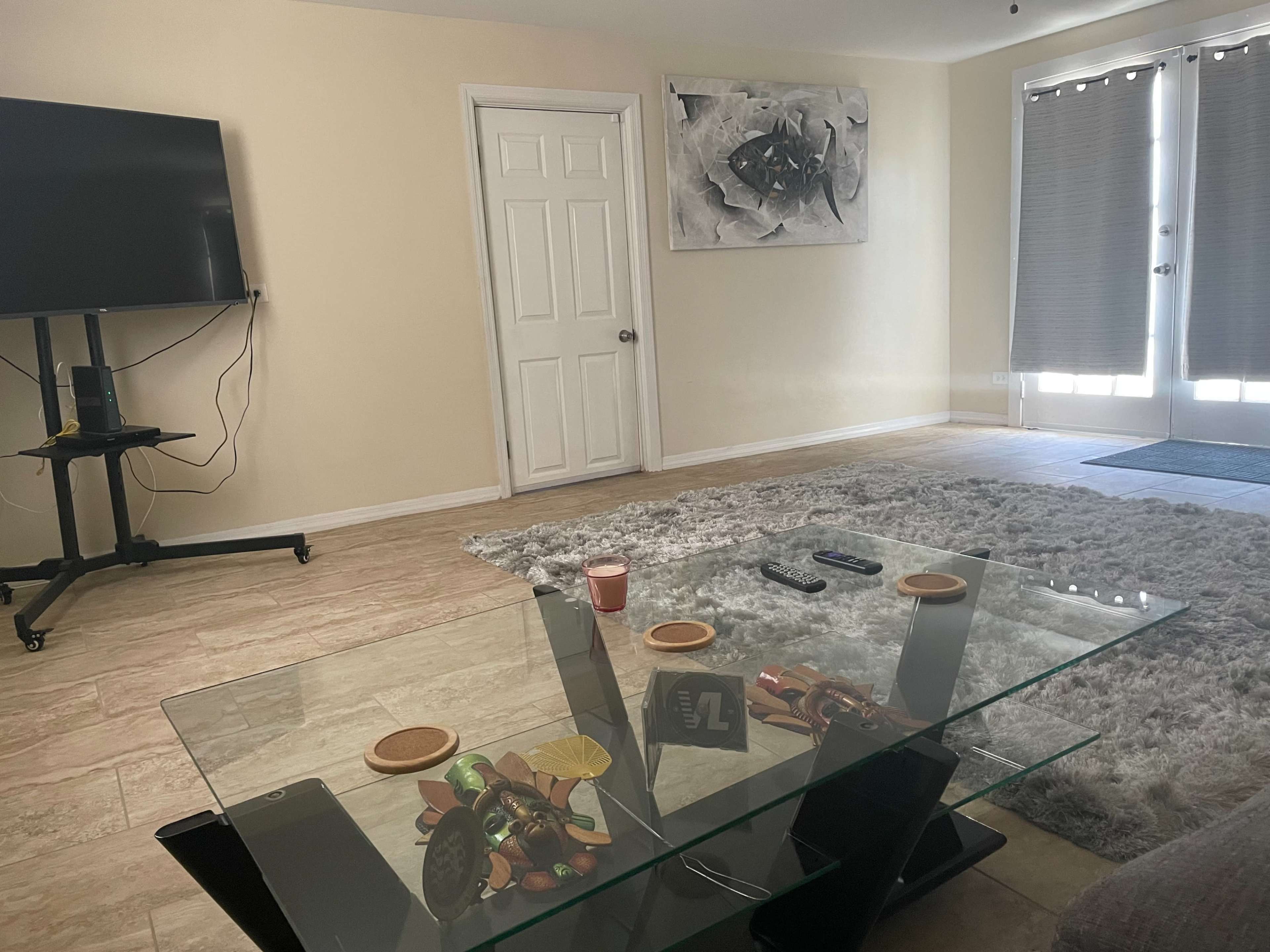 A living room with a glass coffee table, a large gray rug, a TV on a stand, a closed door, and sliding glass doors with blinds.