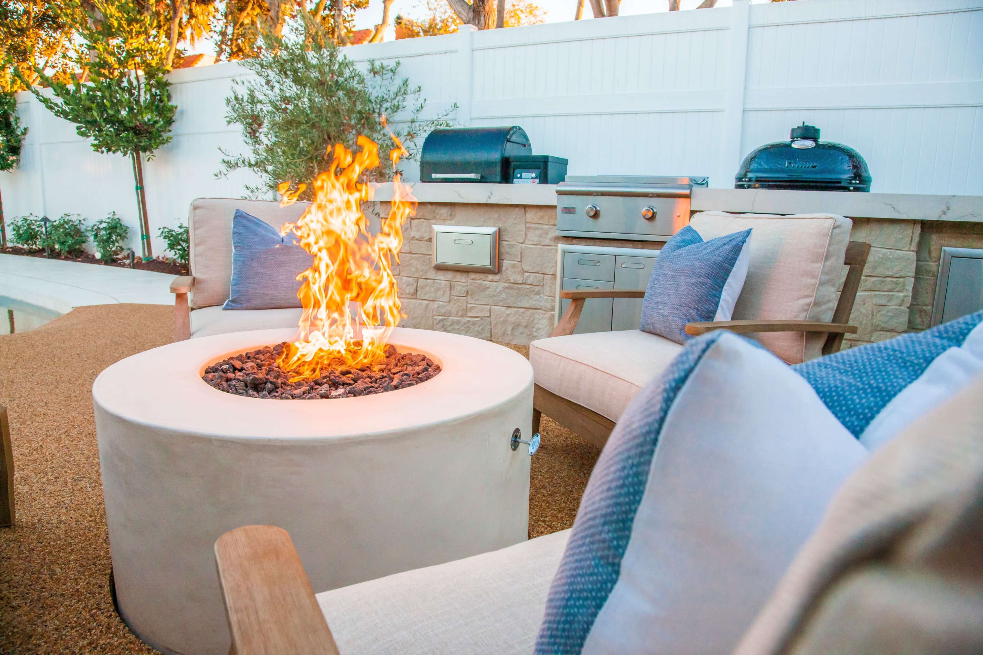 A cozy outdoor seating area features a circular fire pit surrounded by two armchairs, with a barbecue grill visible in the background.