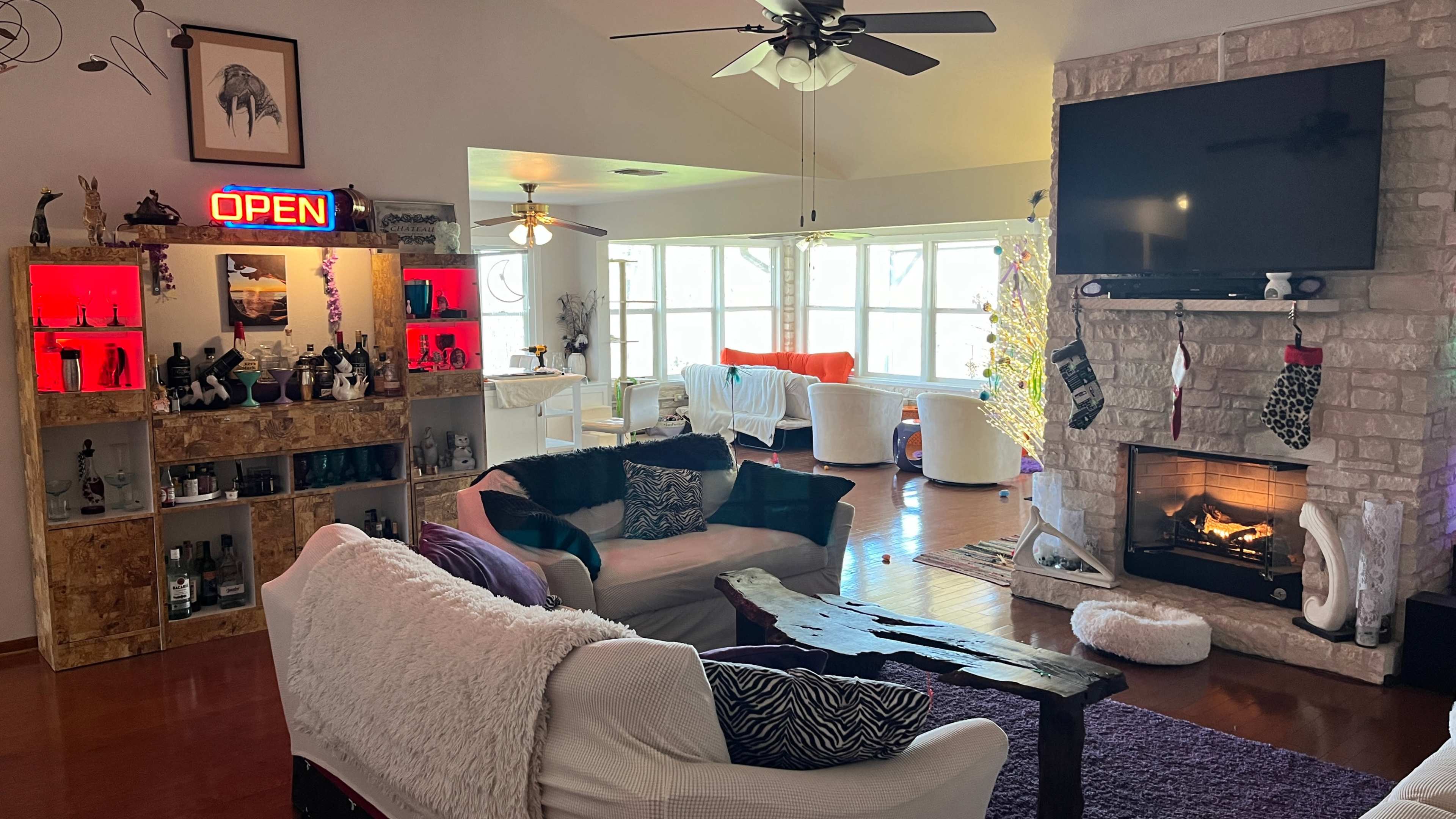 The living room features a cozy arrangement with a fireplace, an "OPEN" neon sign, a wooden coffee table, and a wall-mounted television, surrounded by large windows.