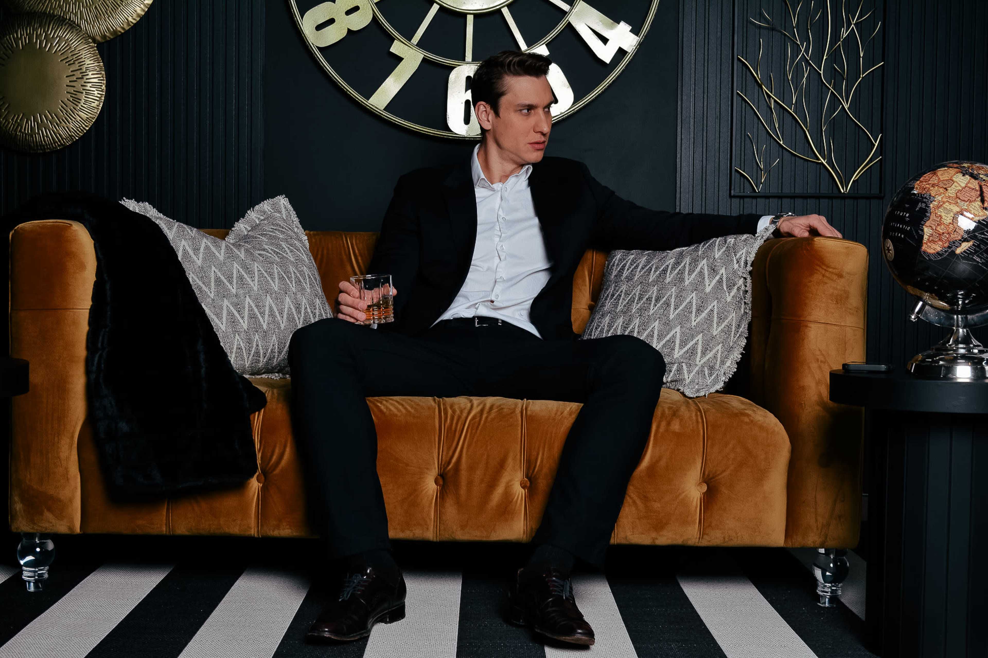 A man in a suit sits on a brown sofa, holding a glass, in a room with a large wall clock and decorative elements.
