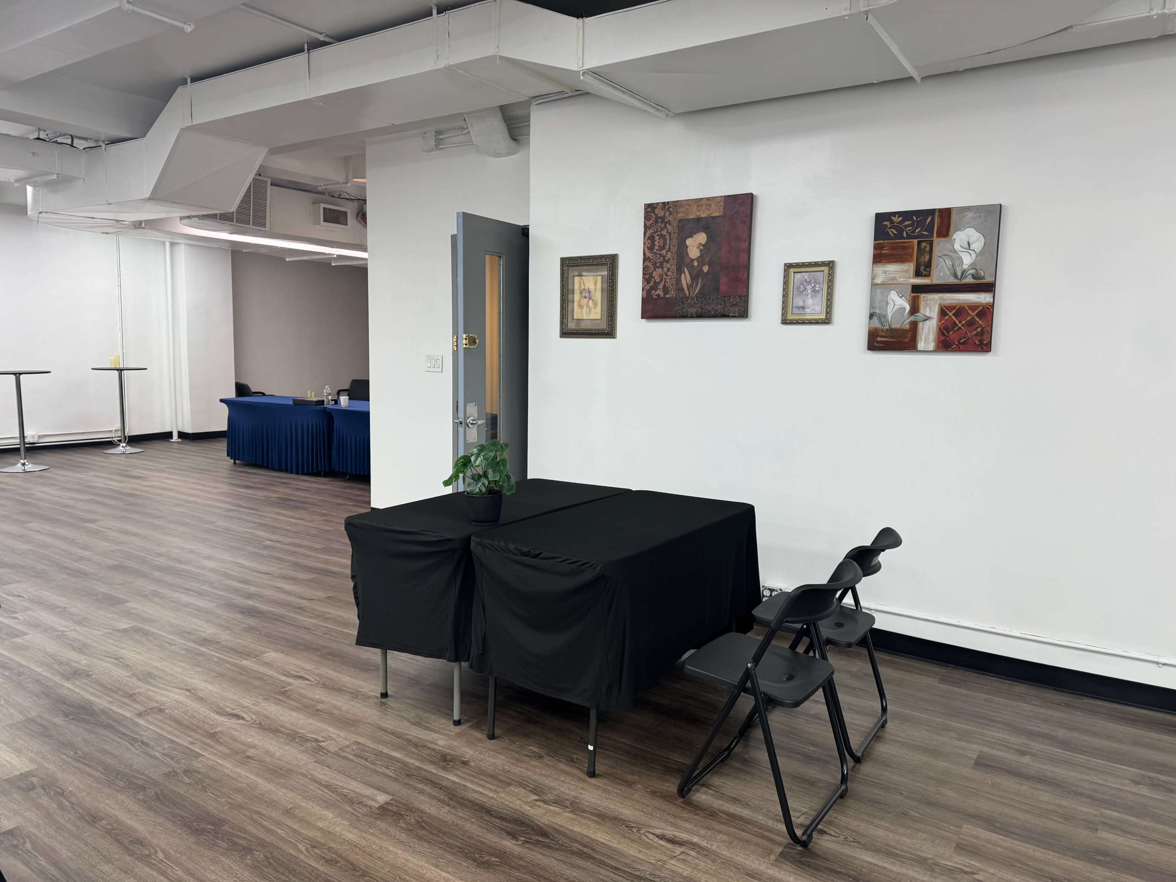The image shows a spacious room with wooden floors, featuring a black table covered with a cloth, two chairs, and framed artwork on the walls.