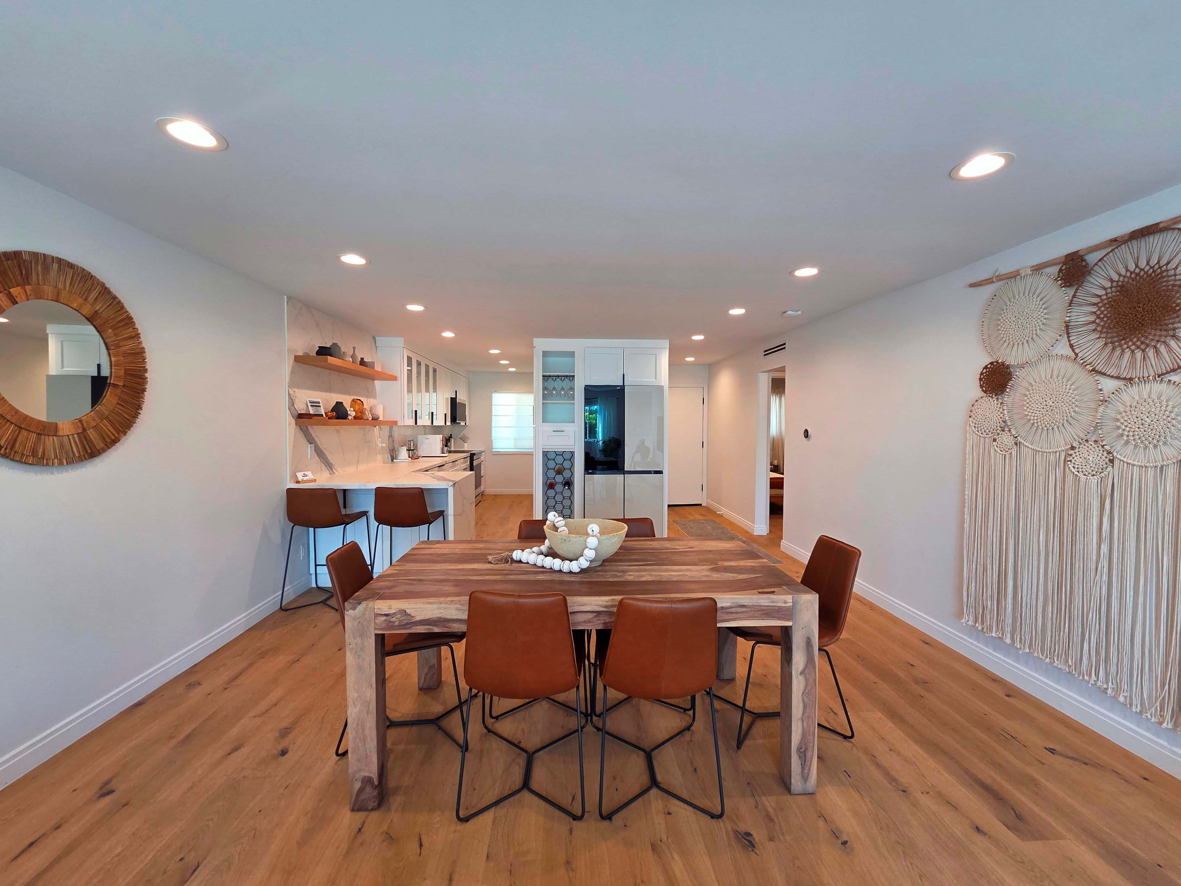 The image depicts a modern dining area with a wooden table surrounded by six brown chairs, adjacent to an open kitchen featuring sleek cabinetry and a large window.