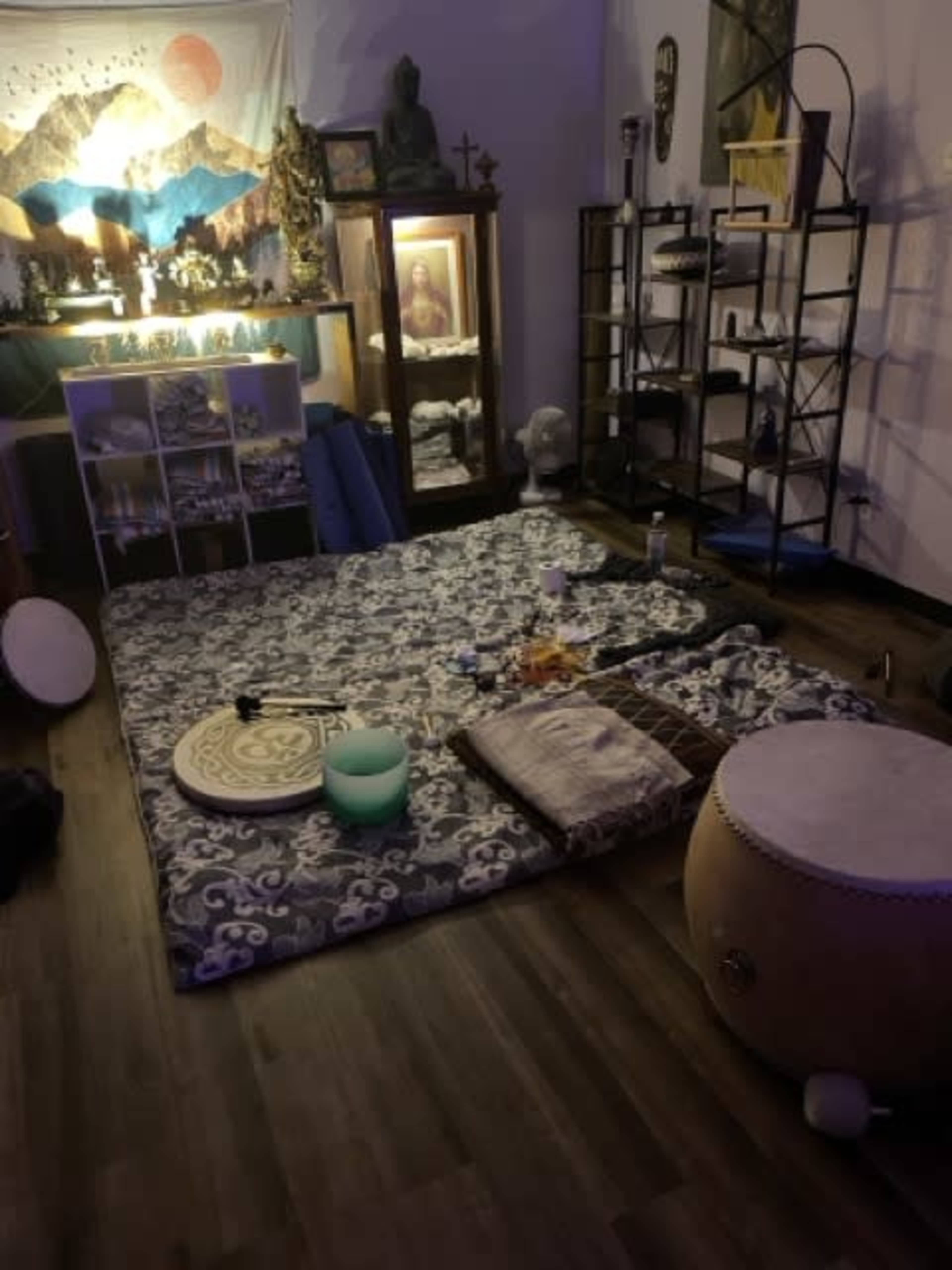 The image shows a meditation space with a large mat in the center, surrounded by various instruments, candles, and decor, including a tapestry depicting mountains and a figure in a frame.