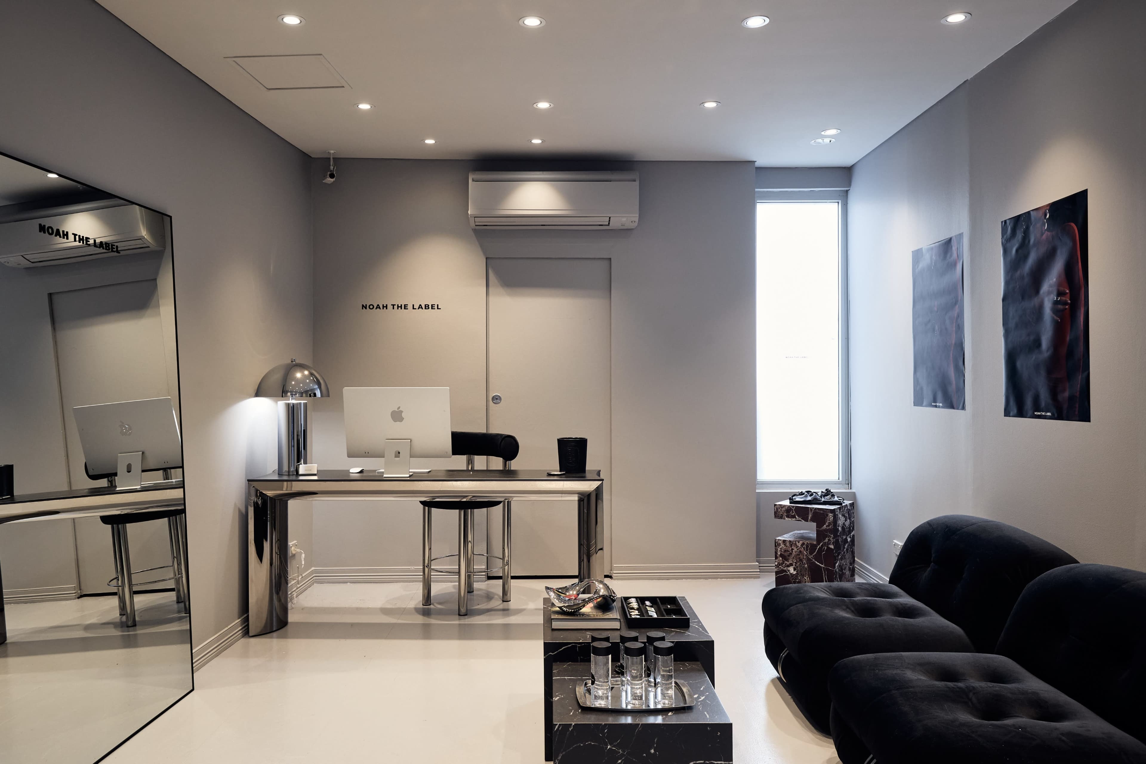 The image shows a modern office space featuring a sleek silver desk with an iMac, a large mirror, black seating, and minimalist decor.