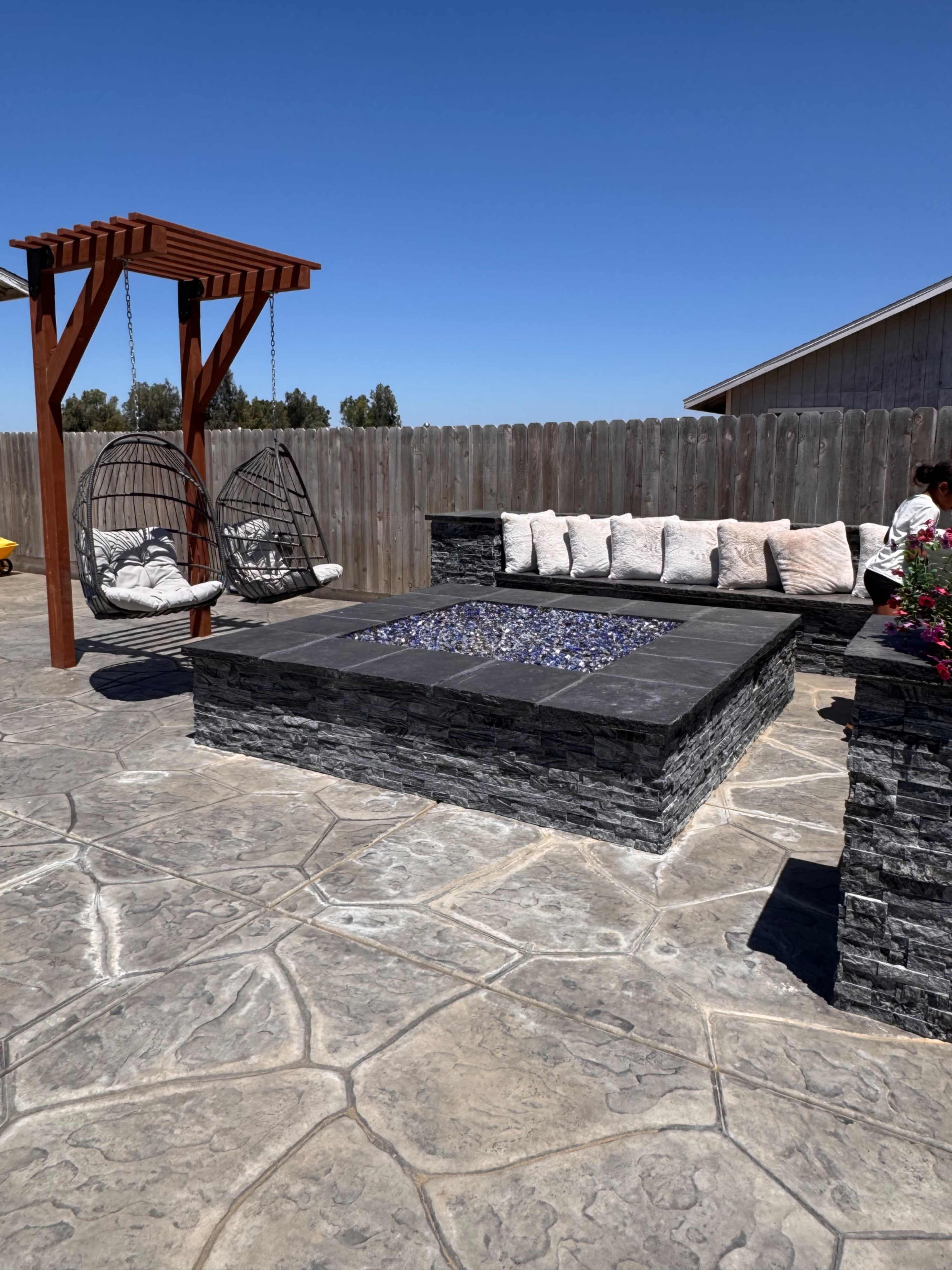 The image shows a patio area featuring a stone fire pit, two hanging swings, and a seating arrangement with decorative pillows.