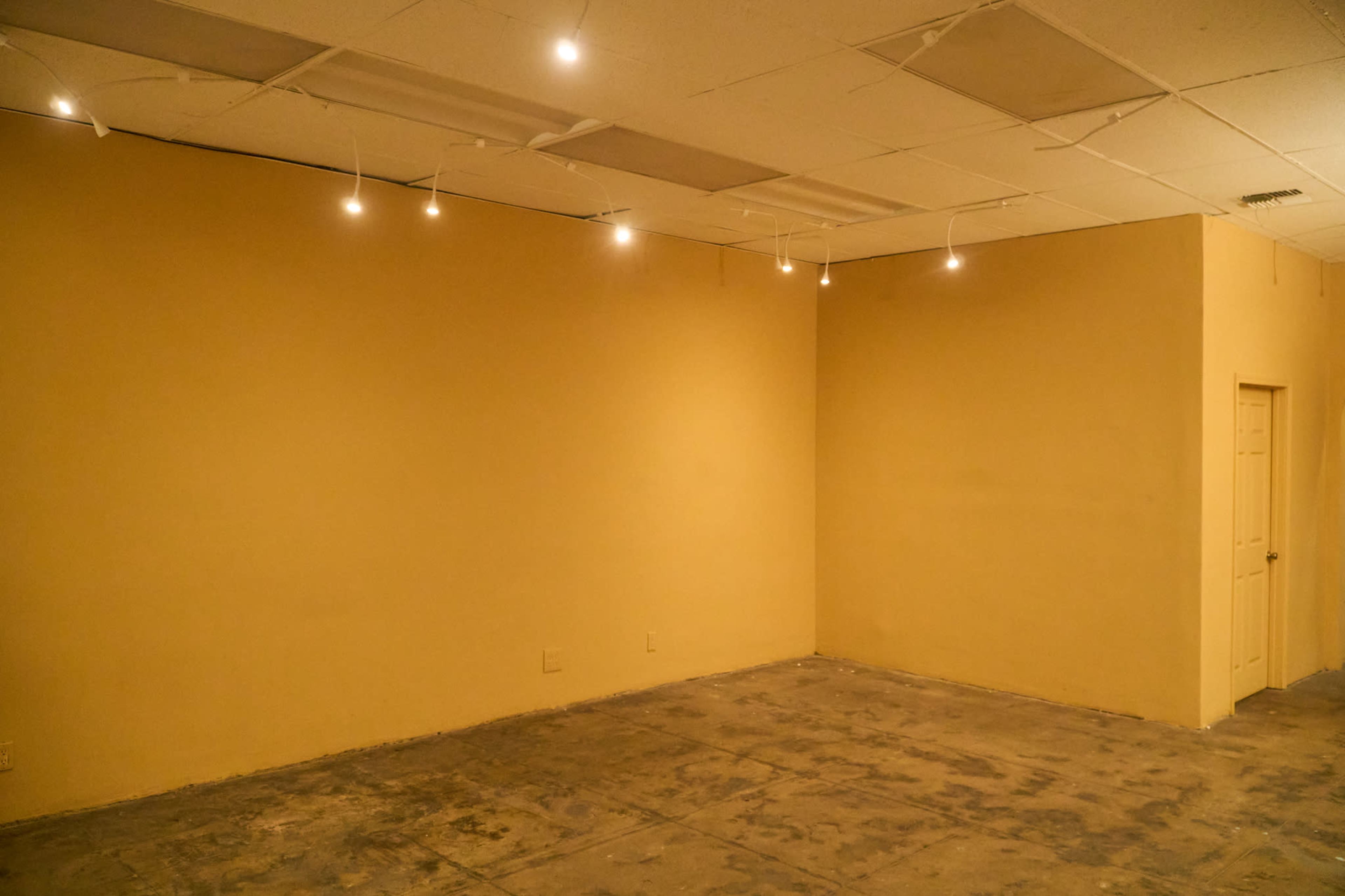 The image shows an empty room with beige walls, a concrete floor, and track lighting on the ceiling.
