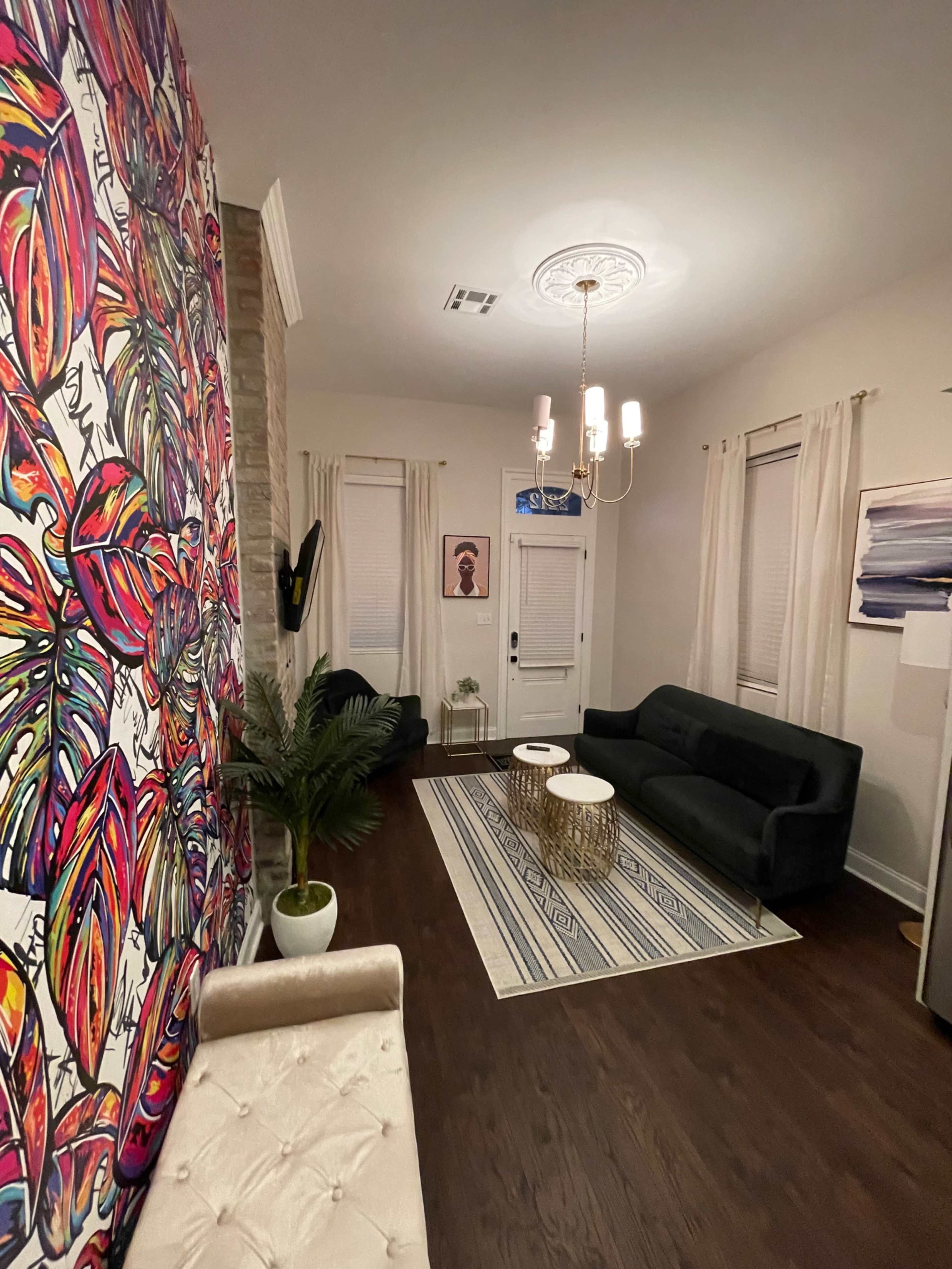The image shows a living room with a colorful floral mural on one wall, dark furniture, a round coffee table, and a door leading outside.