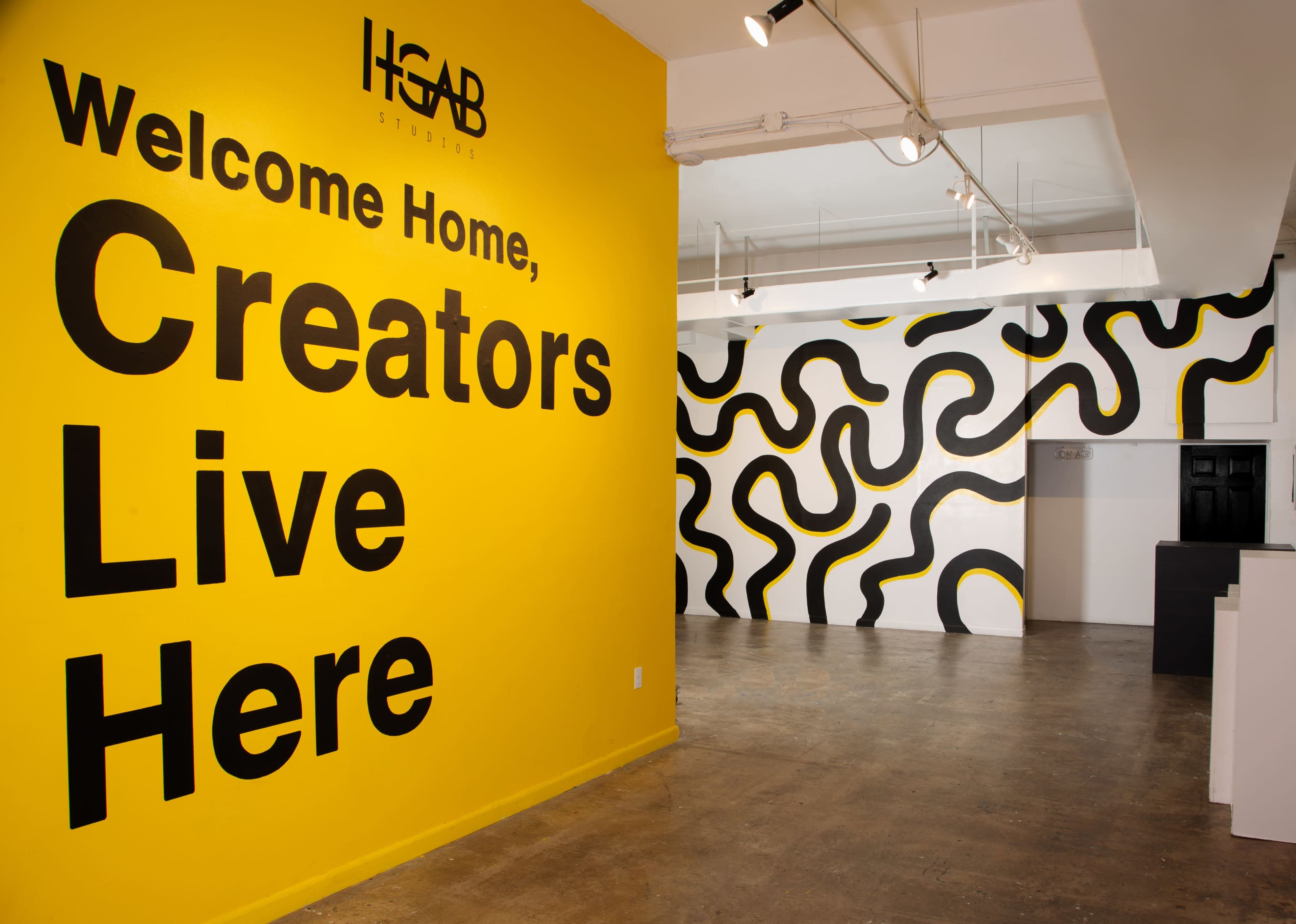A brightly colored yellow wall with large black text stating "Welcome Home, Creators Live Here," alongside a patterned black and yellow mural on the adjacent wall in a spacious studio setting.