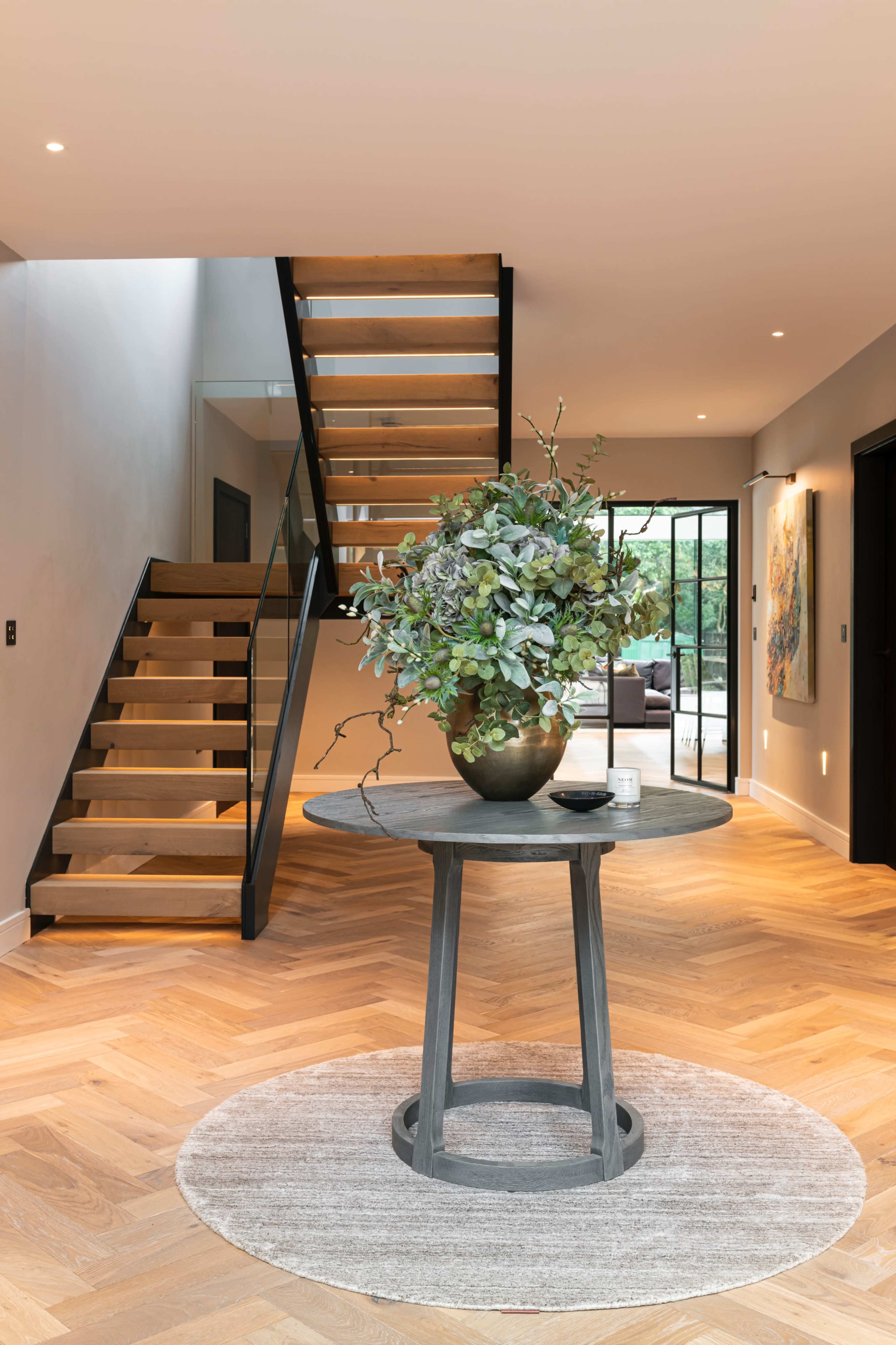 A modern entryway featuring a circular table with a floral arrangement, a staircase with wooden steps, and a rug on a hardwood floor.