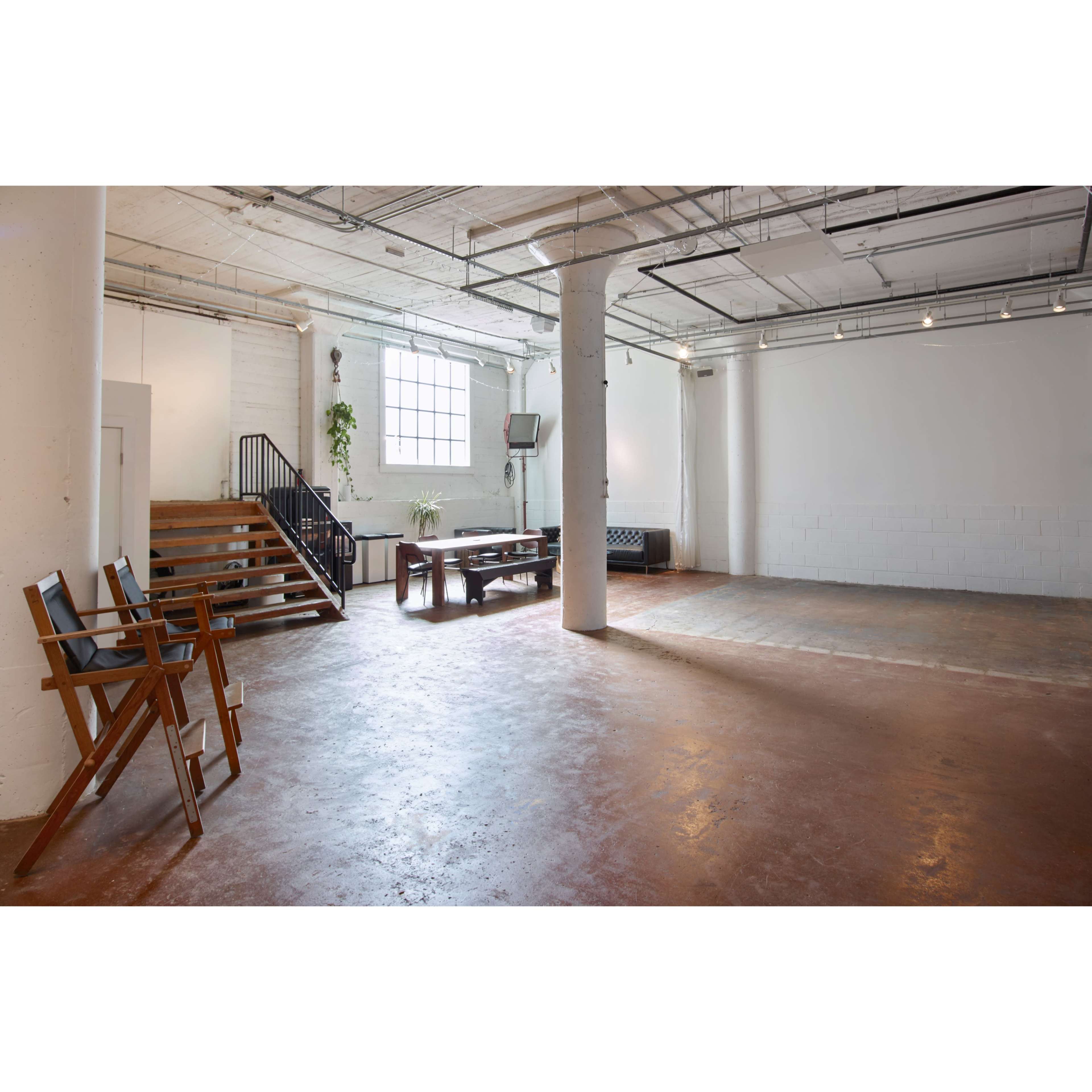 The image shows a spacious, industrial-style room with concrete flooring, a staircase, a dining table, and large windows.