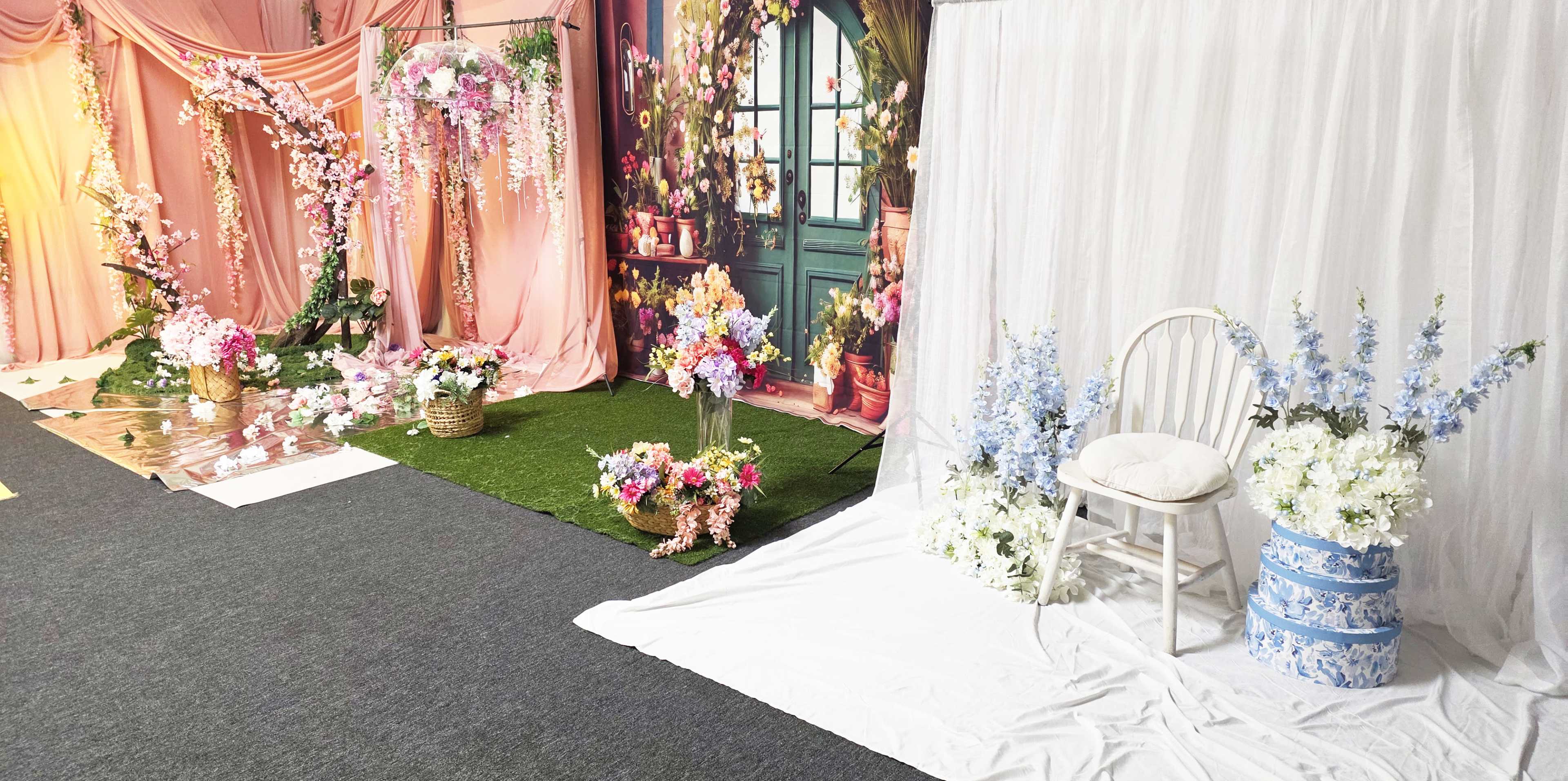 The image shows a decorative space featuring floral arrangements, draped fabric in pastel colors, and a white chair beside blue floral decorations.
