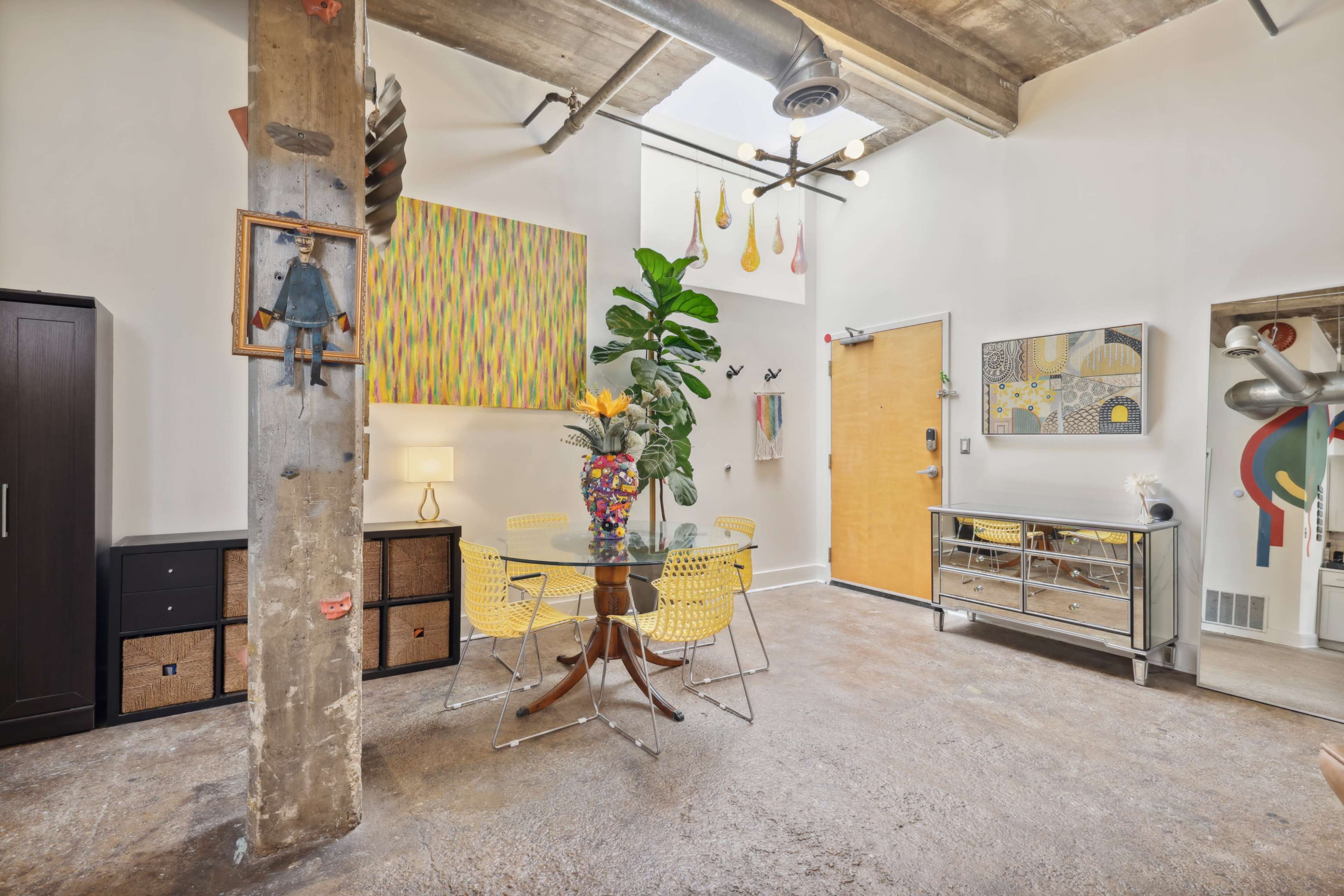 A modern interior space with a dining area featuring a round table, yellow chairs, and vibrant wall art, alongside a concrete pillar and various decorative elements.