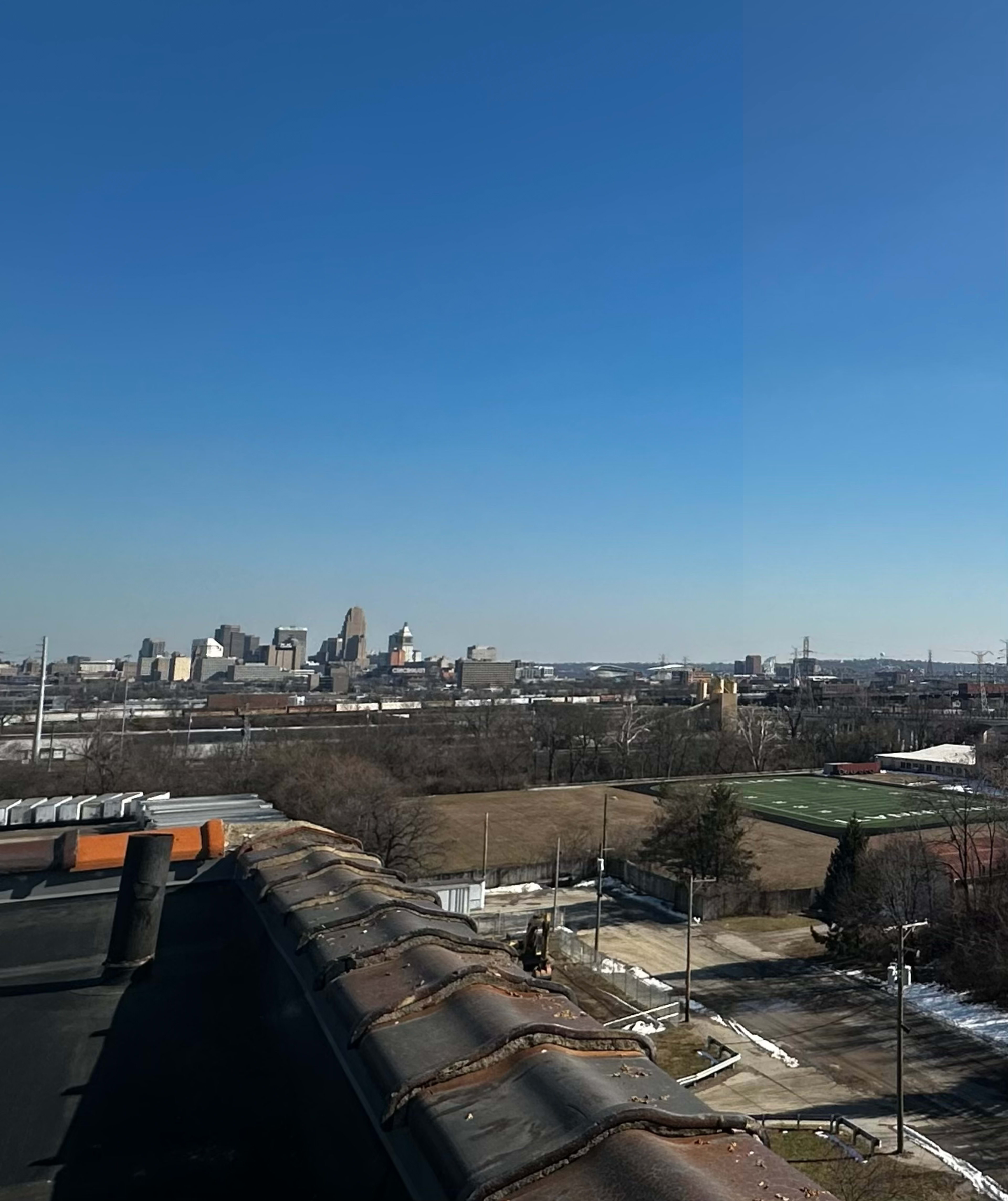 The image shows a view of a city skyline with clear blue skies and a rooftop in the foreground.