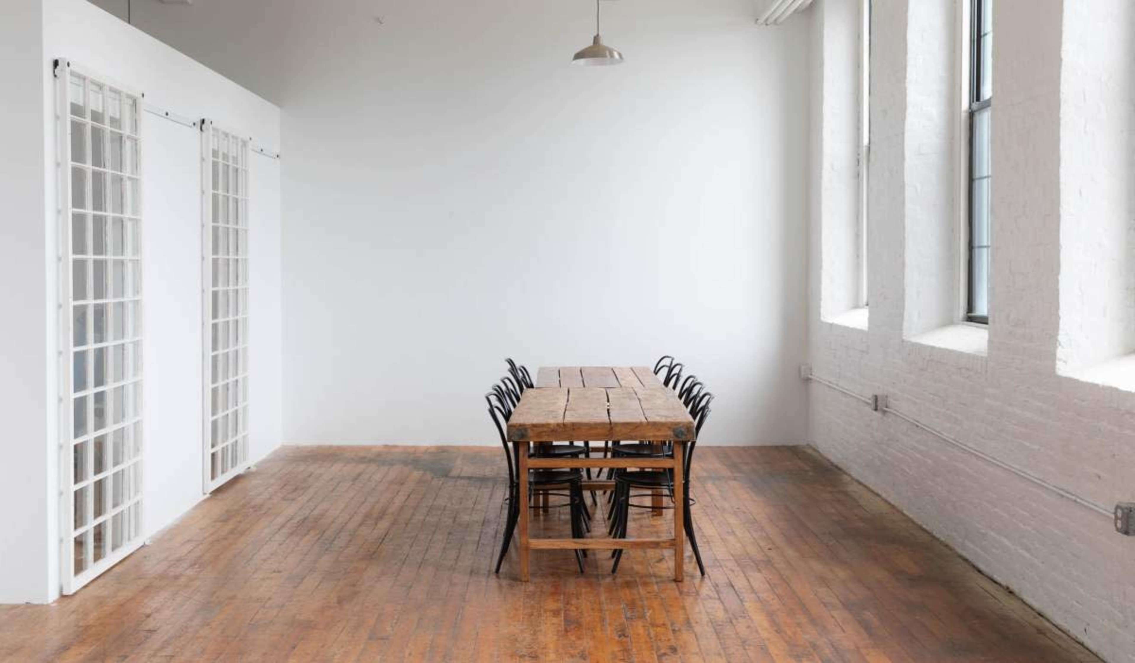 A long wooden table with black chairs is positioned in a bright, minimalist room with large windows and wooden floors.