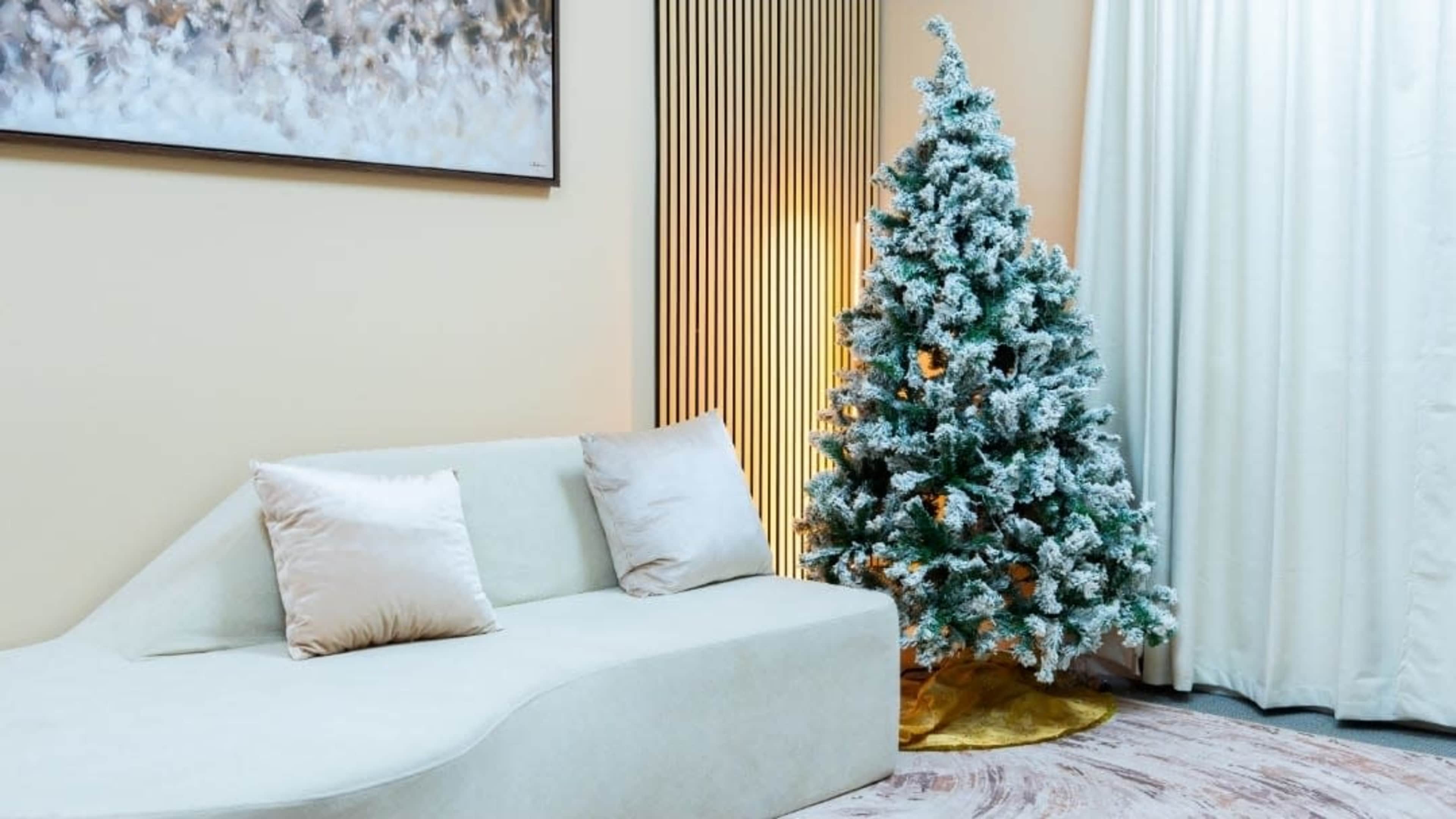 A frosted Christmas tree stands beside a light-colored, curved sofa in a softly lit room with cream-colored walls.