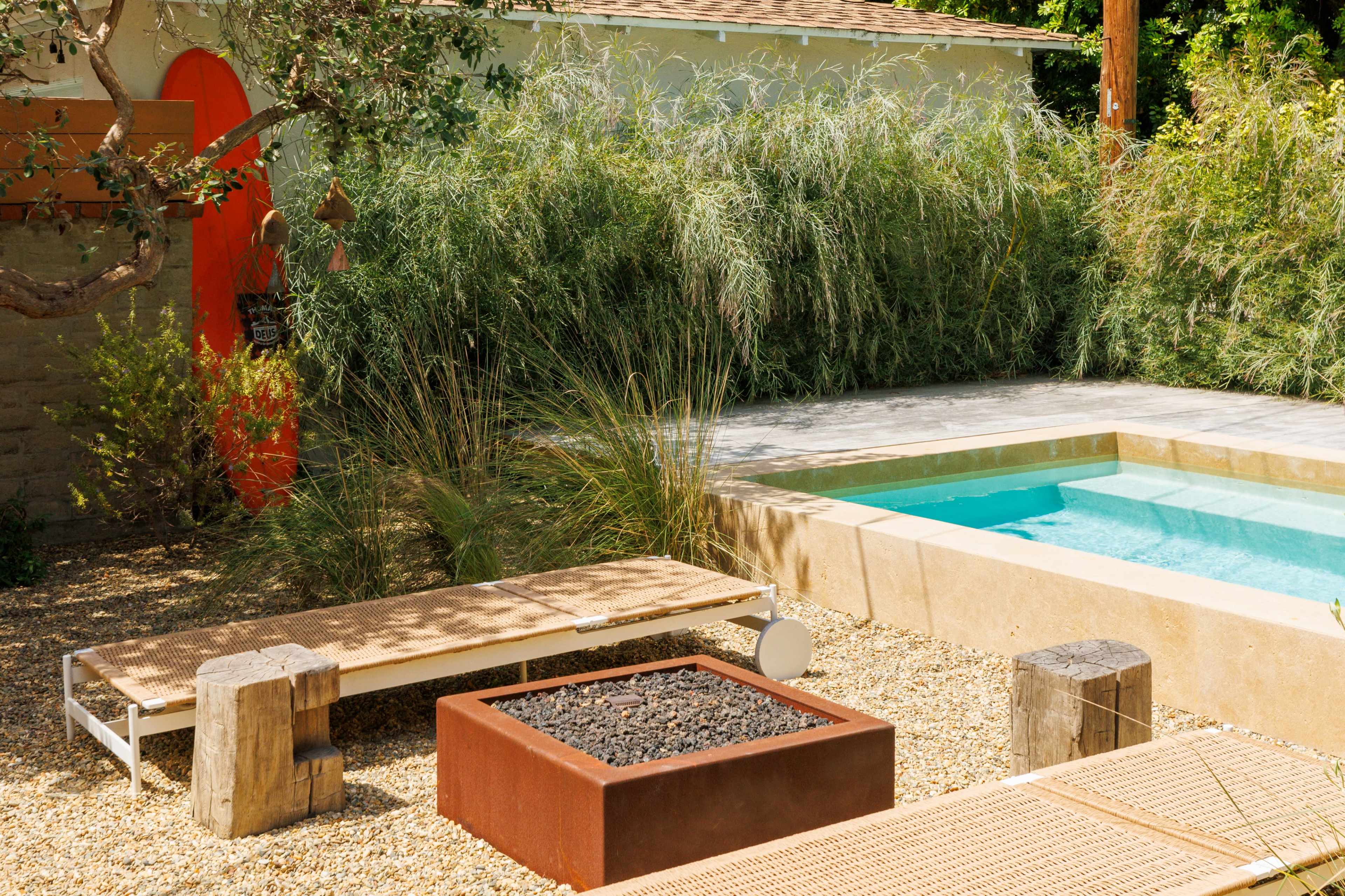 The scene features a landscaped backyard with a swimming pool, a fire pit surrounded by gravel, and lounge chairs near tall grasses and an orange door in the background.