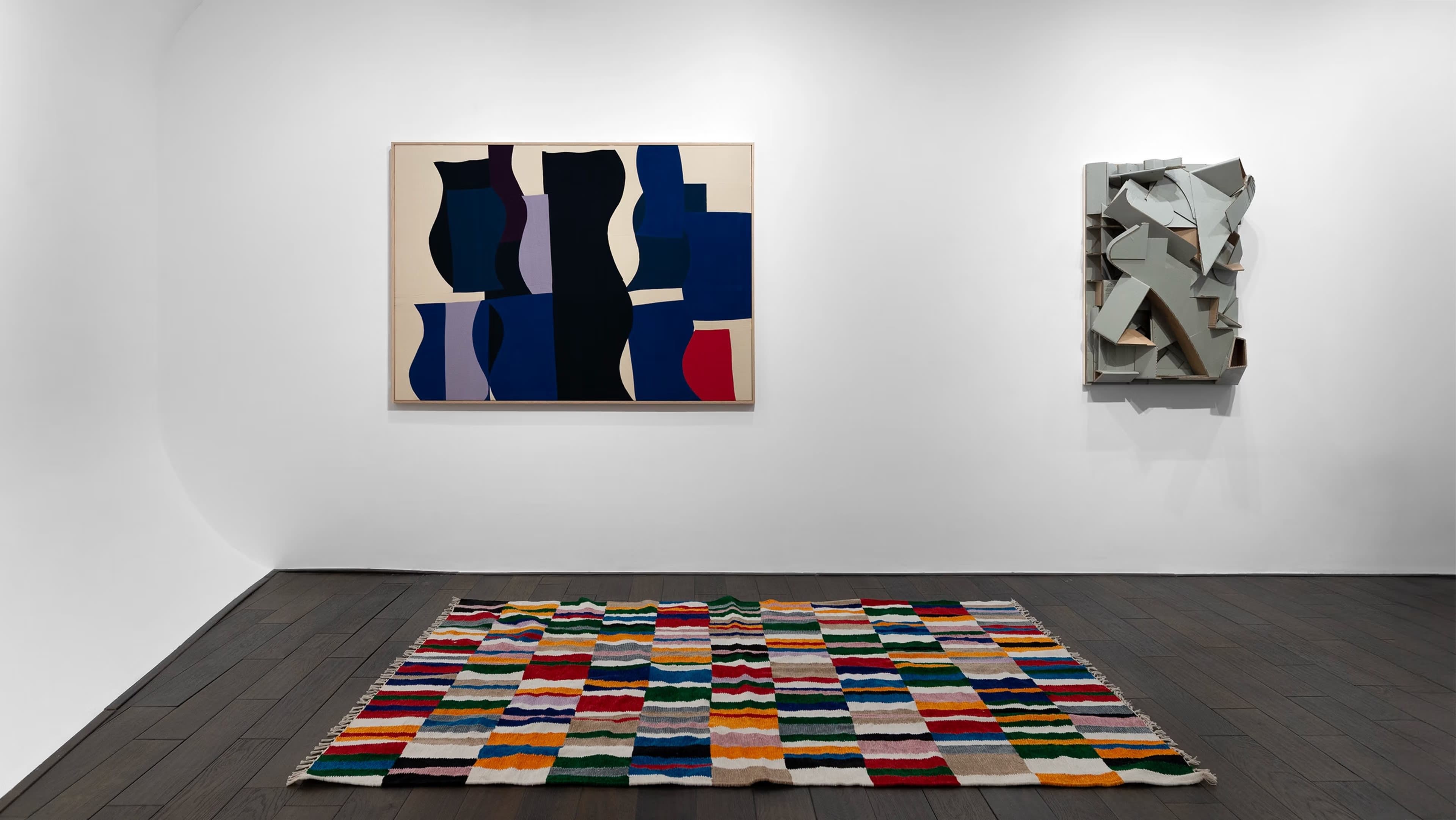 The image shows an art gallery with two abstract paintings on the wall and a colorful geometric rug on the floor.