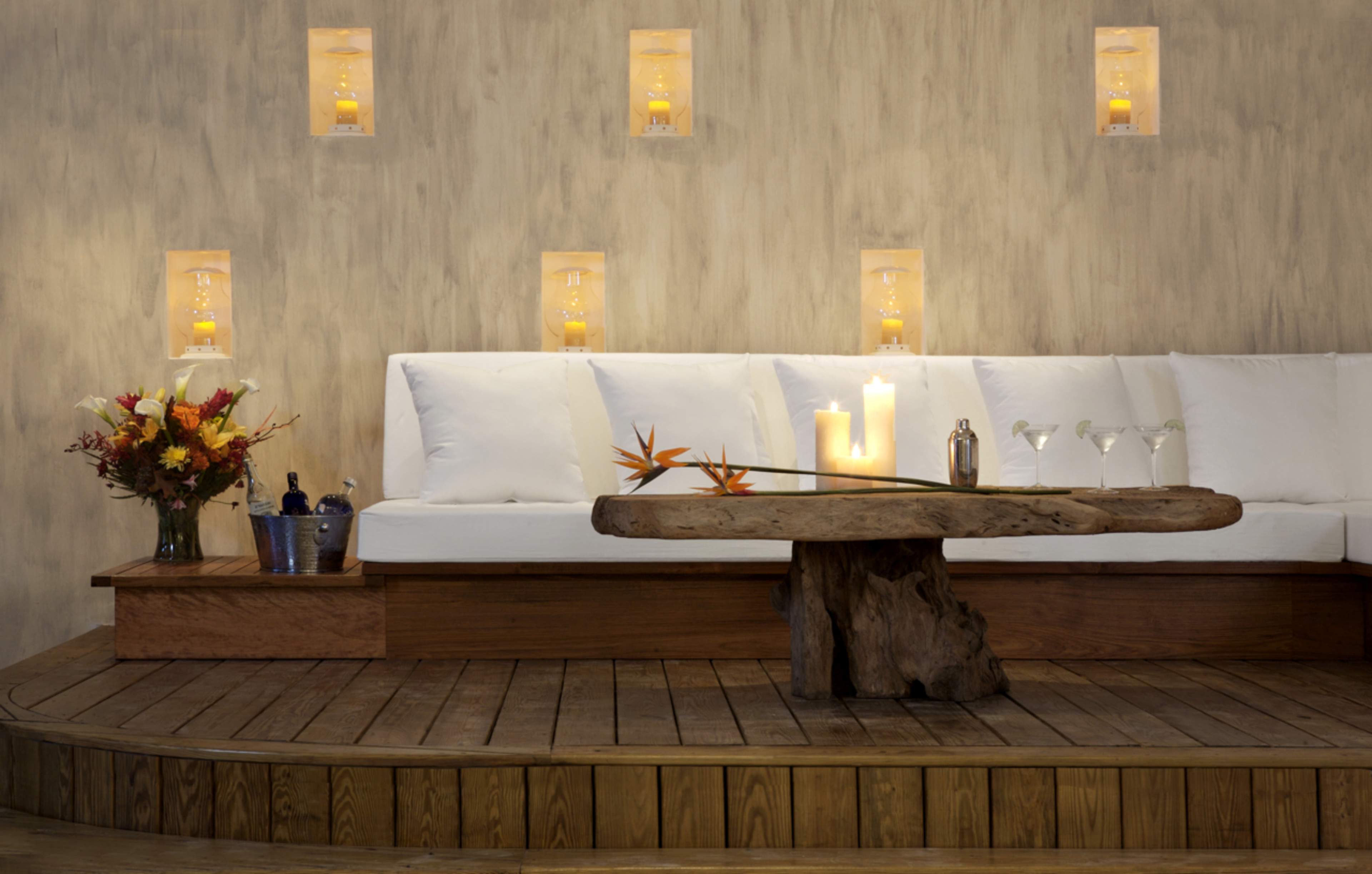 A modern, minimalist lounge space features a wooden table, a white sofa arranged along the wall, and softly glowing candles set in wall alcoves, with a bouquet of flowers on one side.