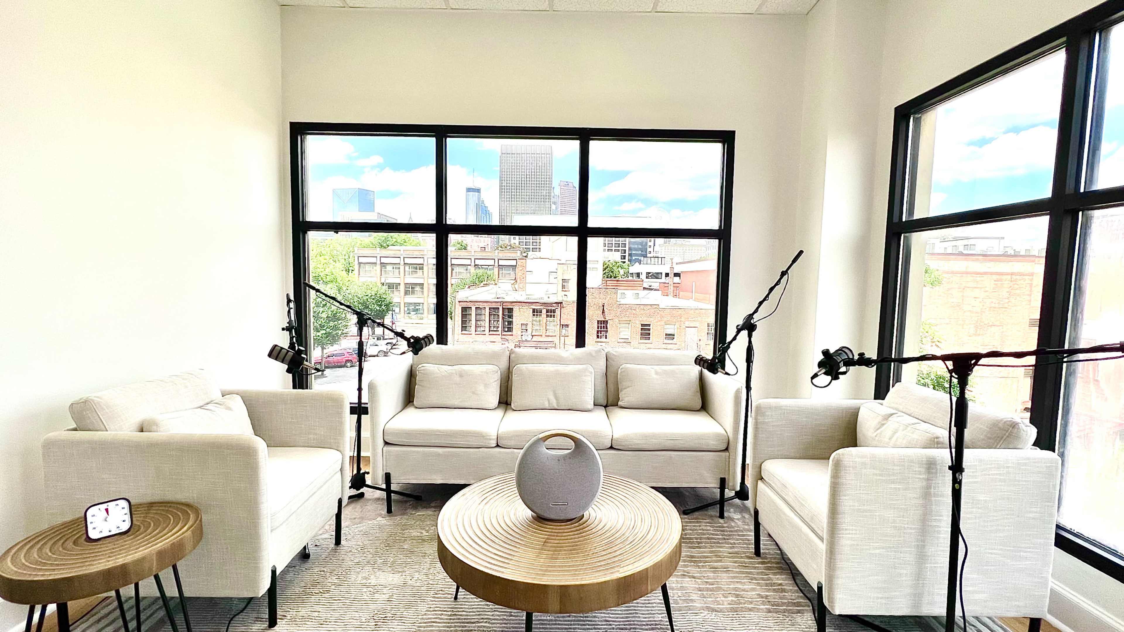 A bright interior features two white sofas and a round coffee table near large windows overlooking a cityscape.