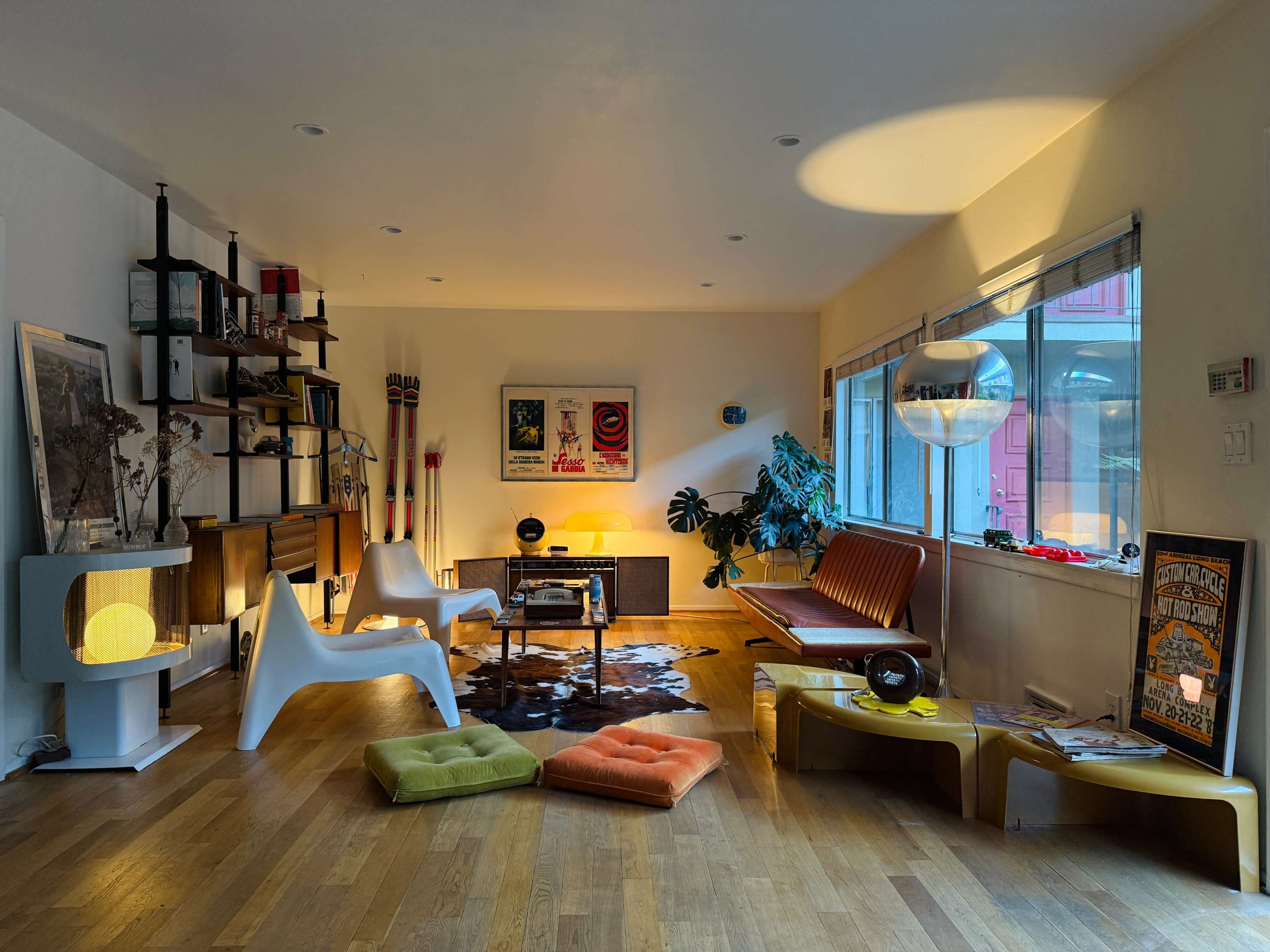 The living room features a mix of mid-century modern furniture, including a leather sofa, colorful cushions, and decorative shelving, all illuminated by natural light.