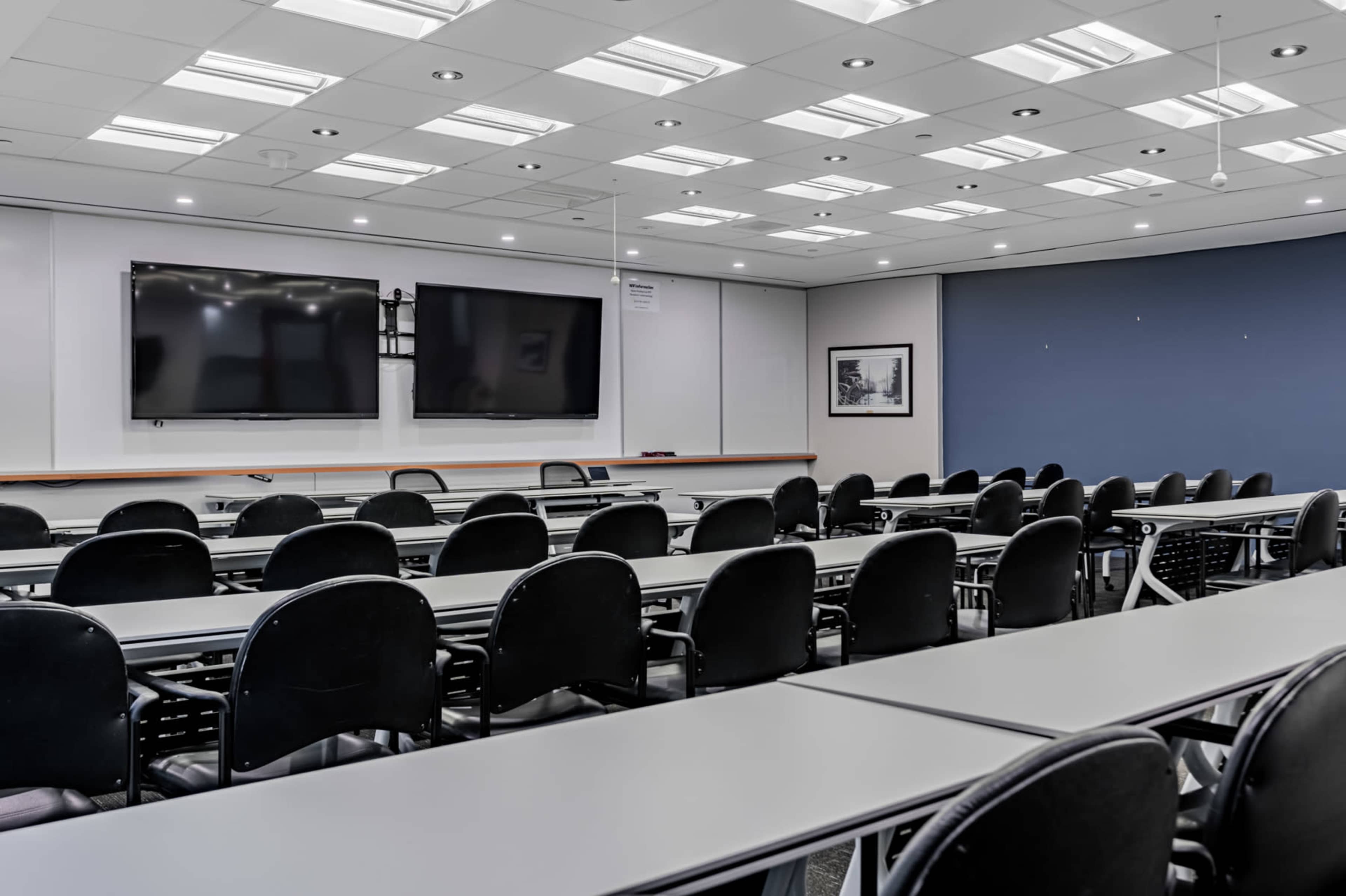 A modern classroom setup with rows of black chairs and desks, two wall-mounted screens, and a blue accent wall.