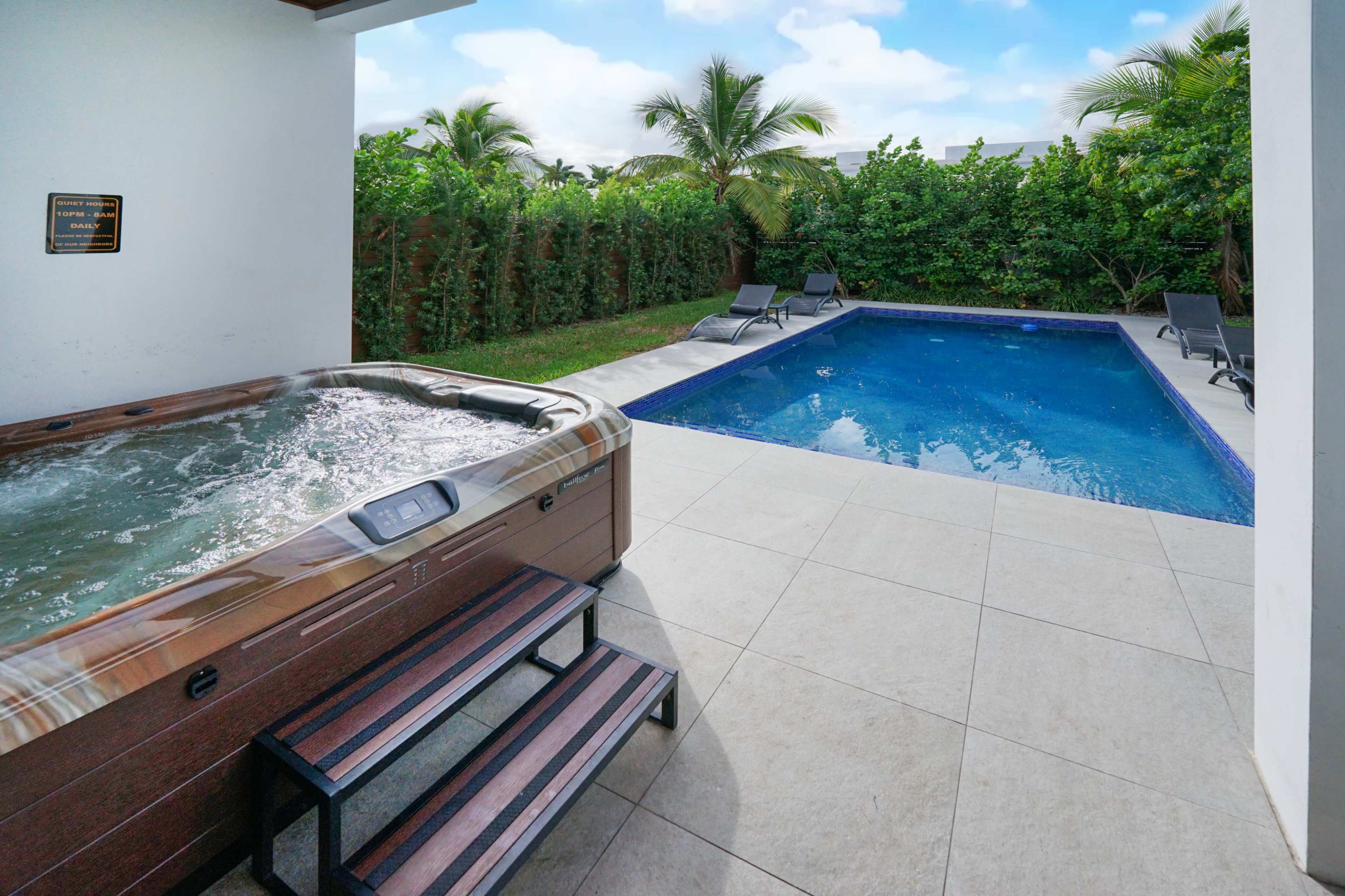 The image shows a backyard with a hot tub and a swimming pool surrounded by palm trees and lounge chairs.