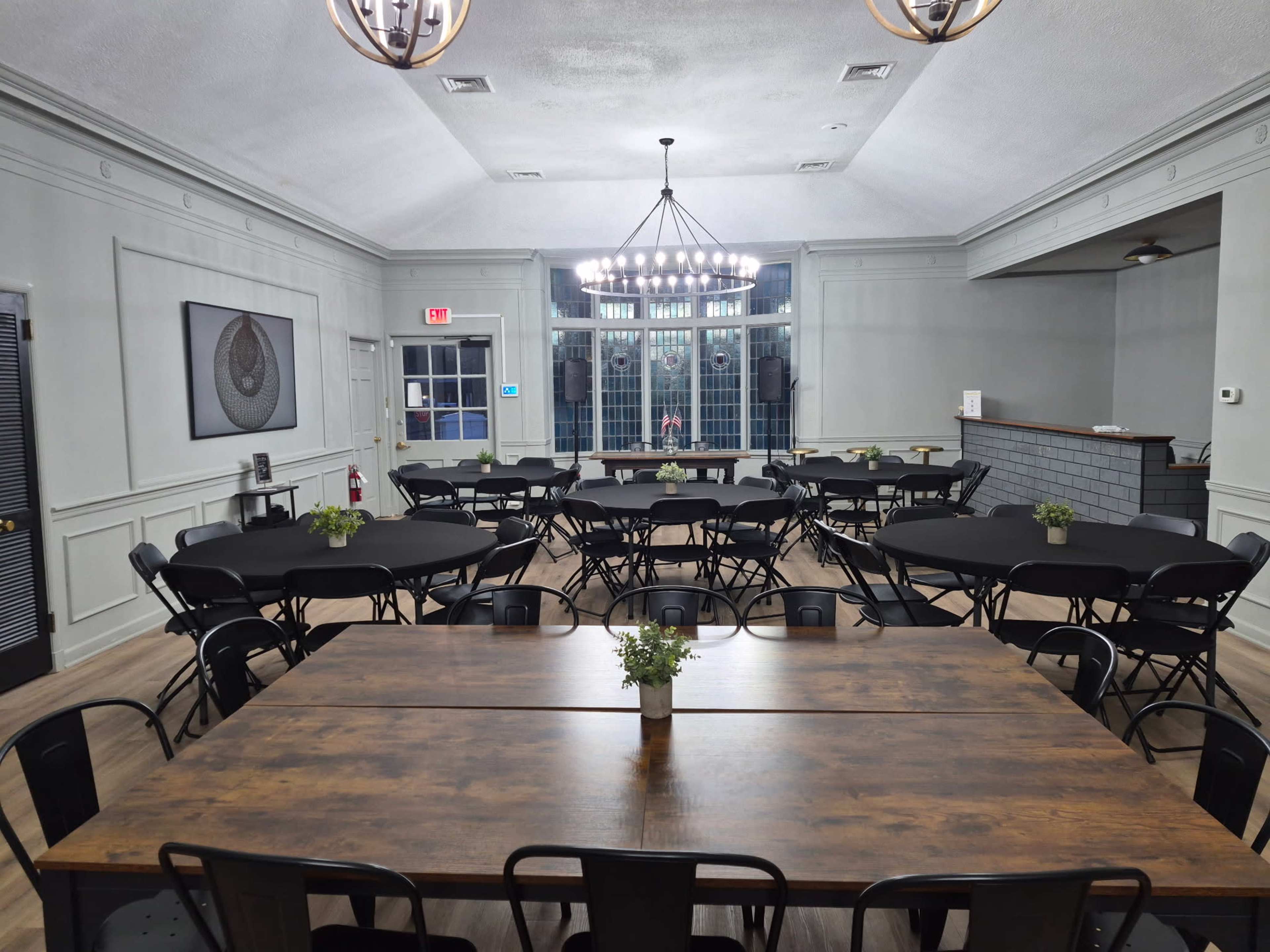 The image shows a spacious event room with multiple round and rectangular tables arranged neatly, featuring black tablecloths and small centerpieces, illuminated by a chandelier and natural light from large windows.