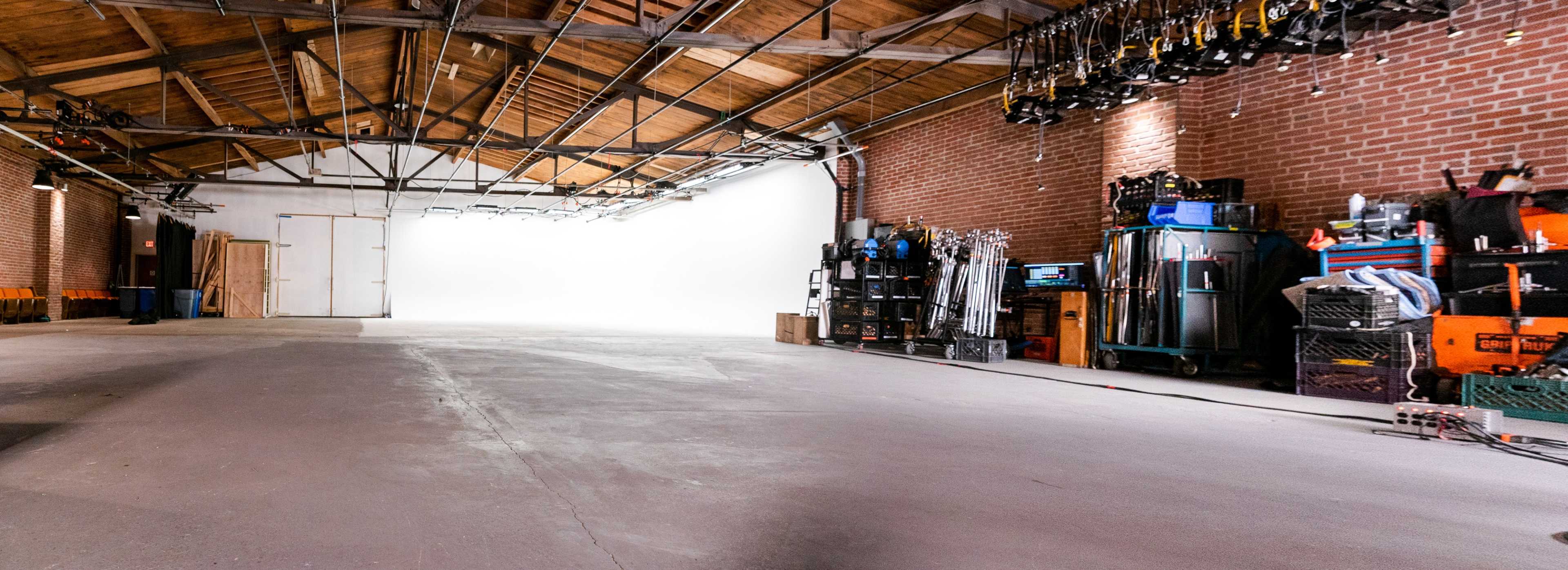 A spacious, empty warehouse with a white backdrop at one end and various equipment neatly stored along the walls.