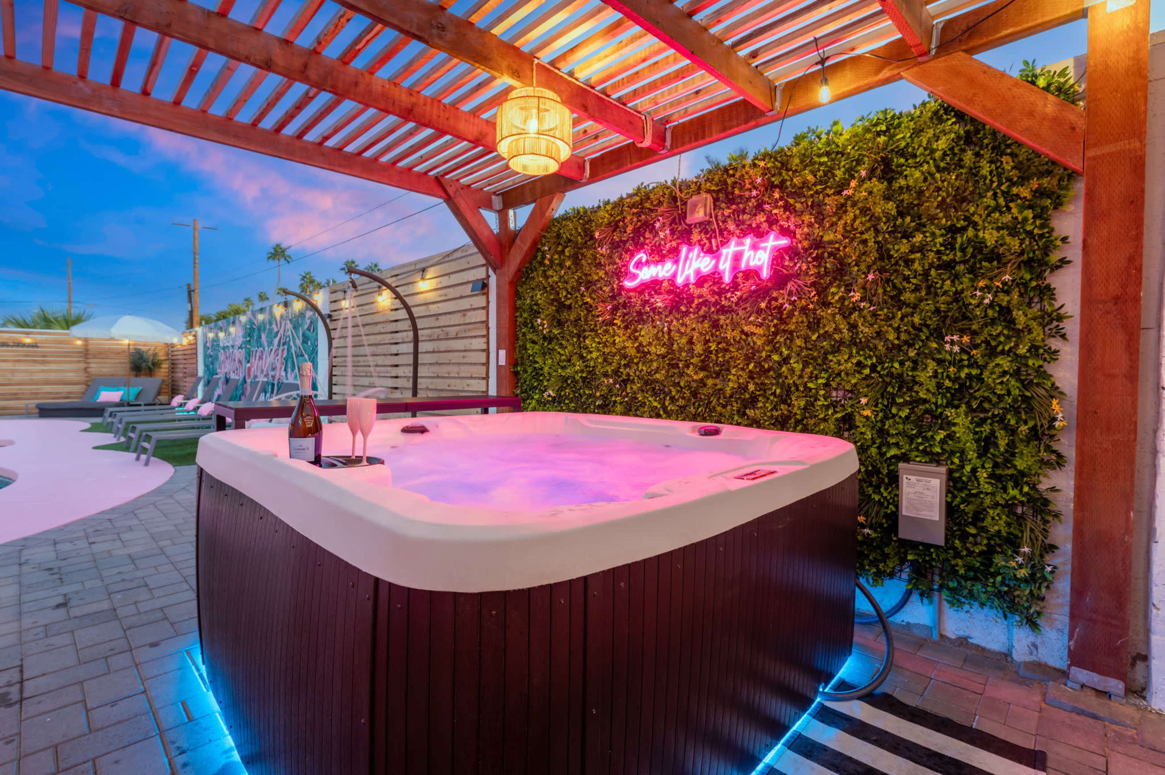 A hot tub with pink lighting is situated under a wooden pergola, next to a green wall with a neon sign, and surrounded by lounge chairs and outdoor decor.
