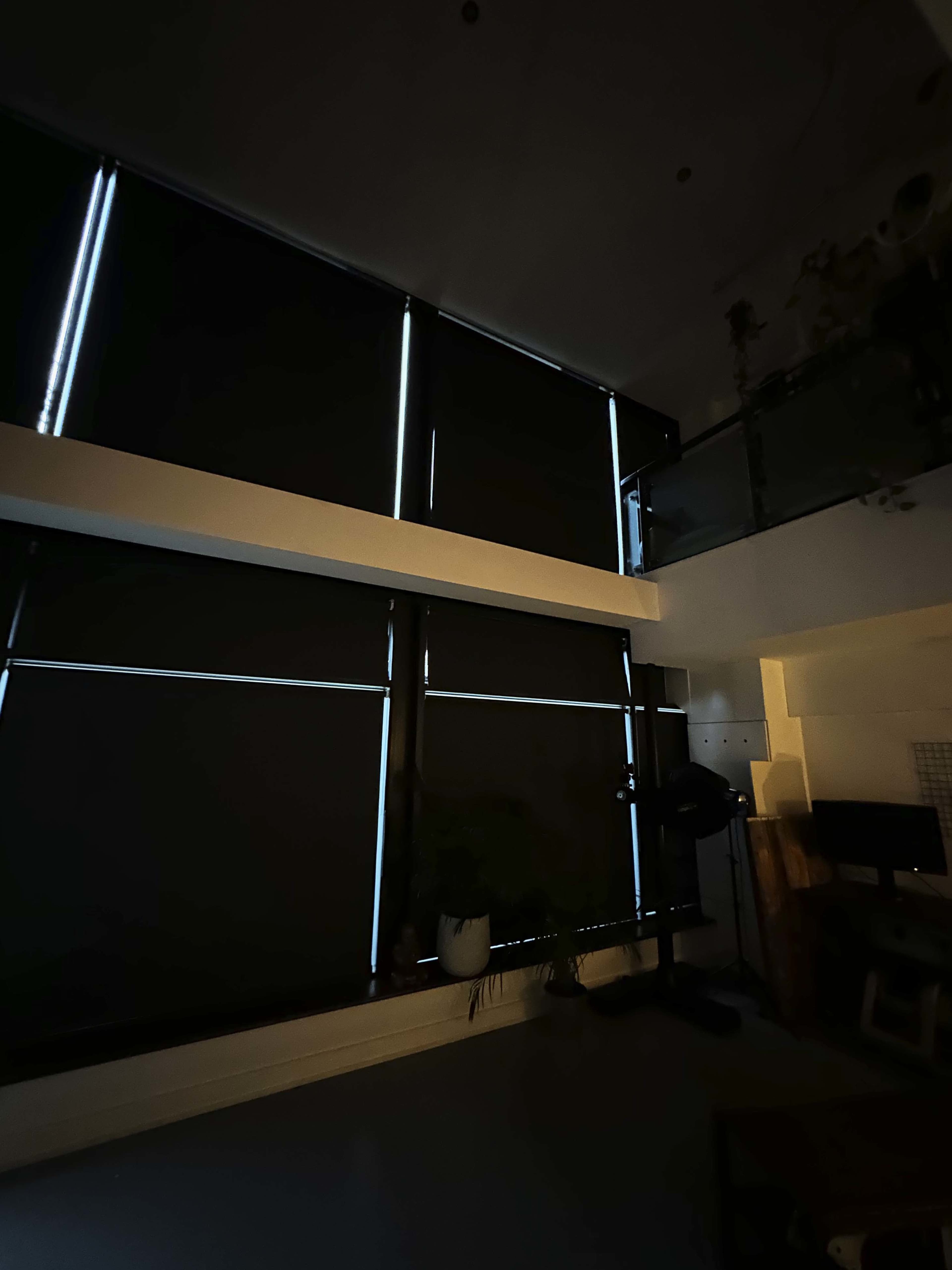 A dimly lit room with large black window blinds partially open, allowing thin streams of light to enter.