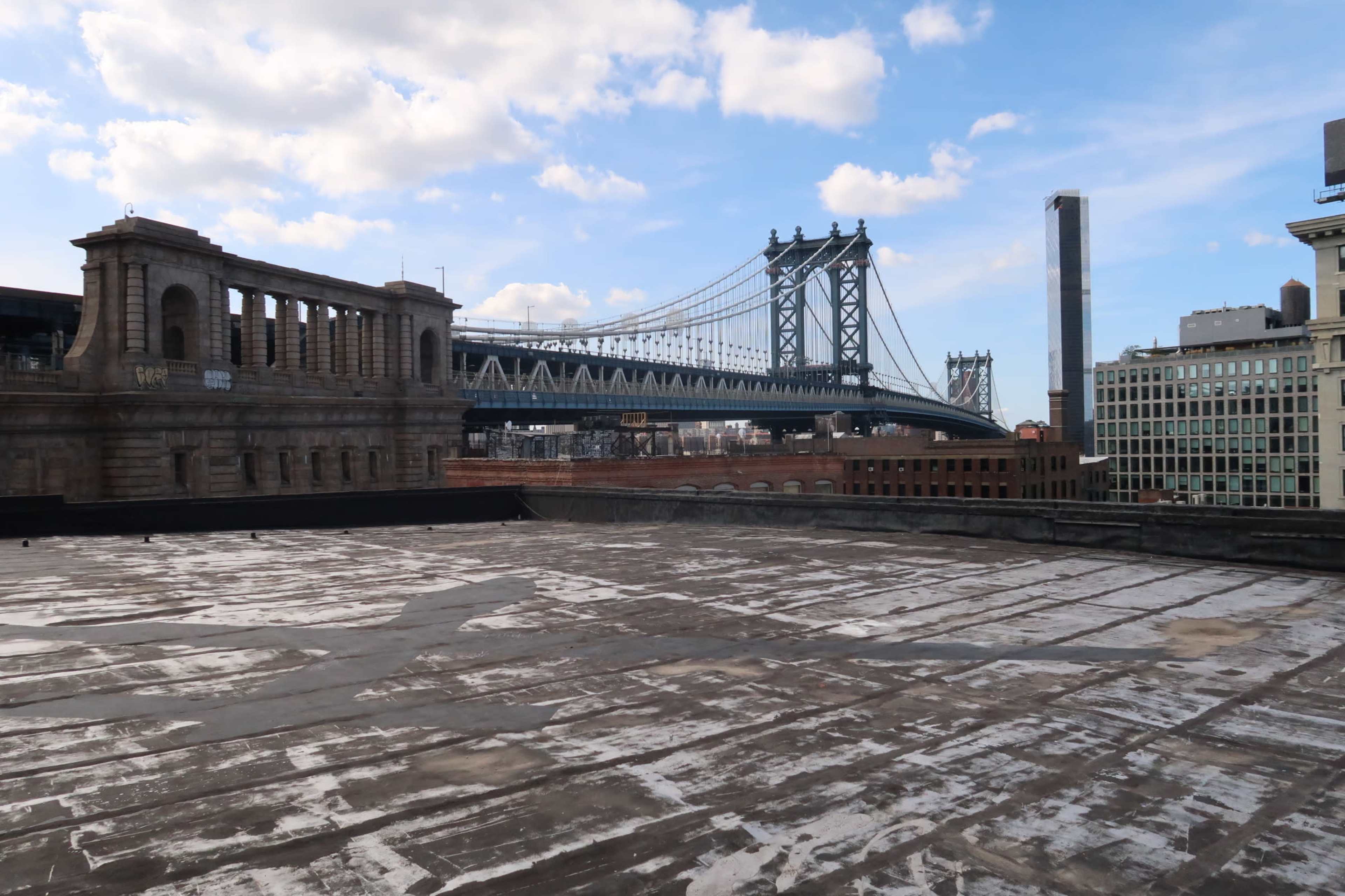 The Manhattan Bridge spanning over a rooftop with a weathered surface, set against a backdrop of blue skies and clouds.
