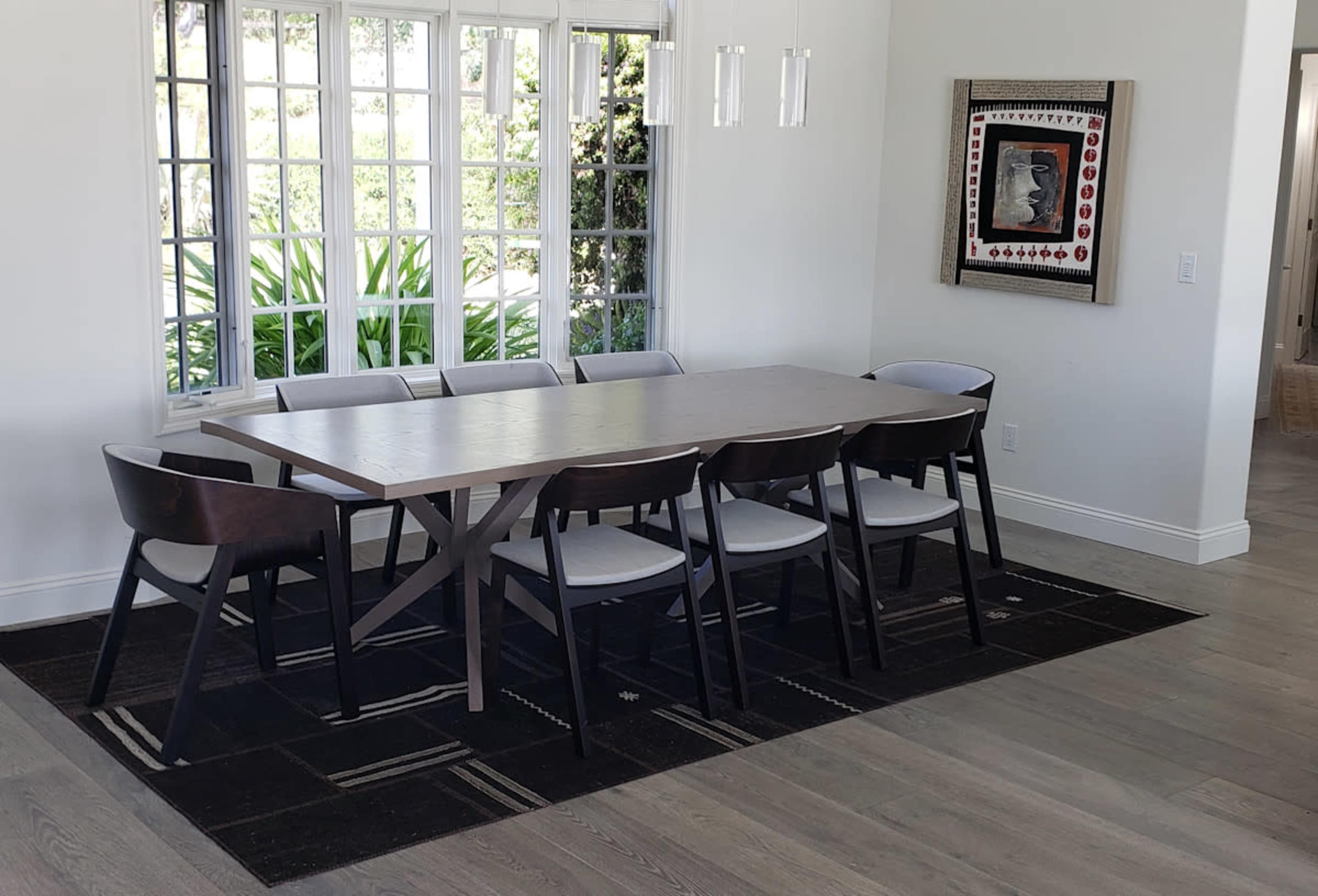 A rectangular dining table with six chairs is set on a patterned rug under hanging light fixtures next to a window with multiple panes.