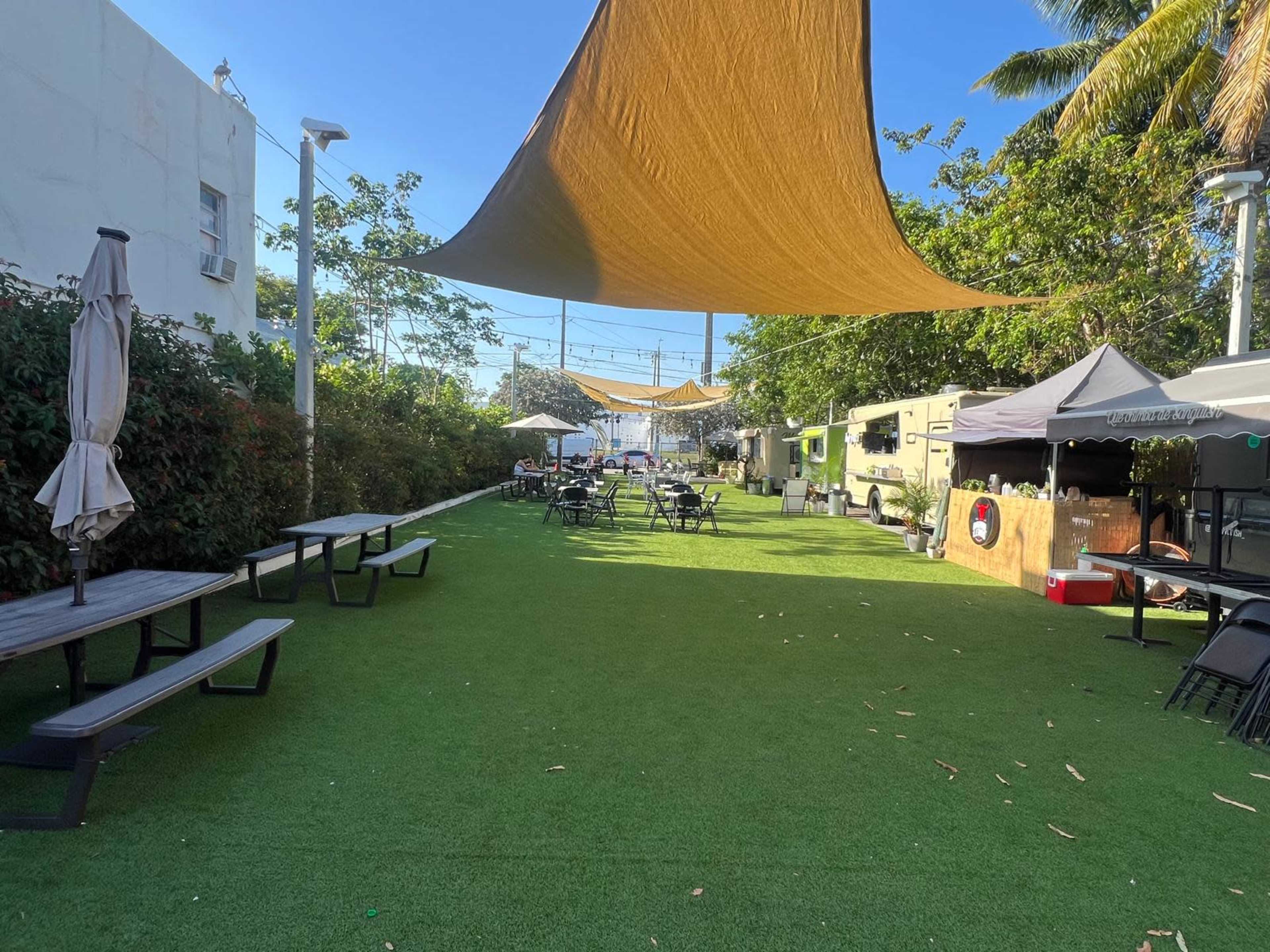 The image shows a spacious outdoor area with artificial grass, shaded by large fabric canopies, featuring tables and chairs set up for seating.