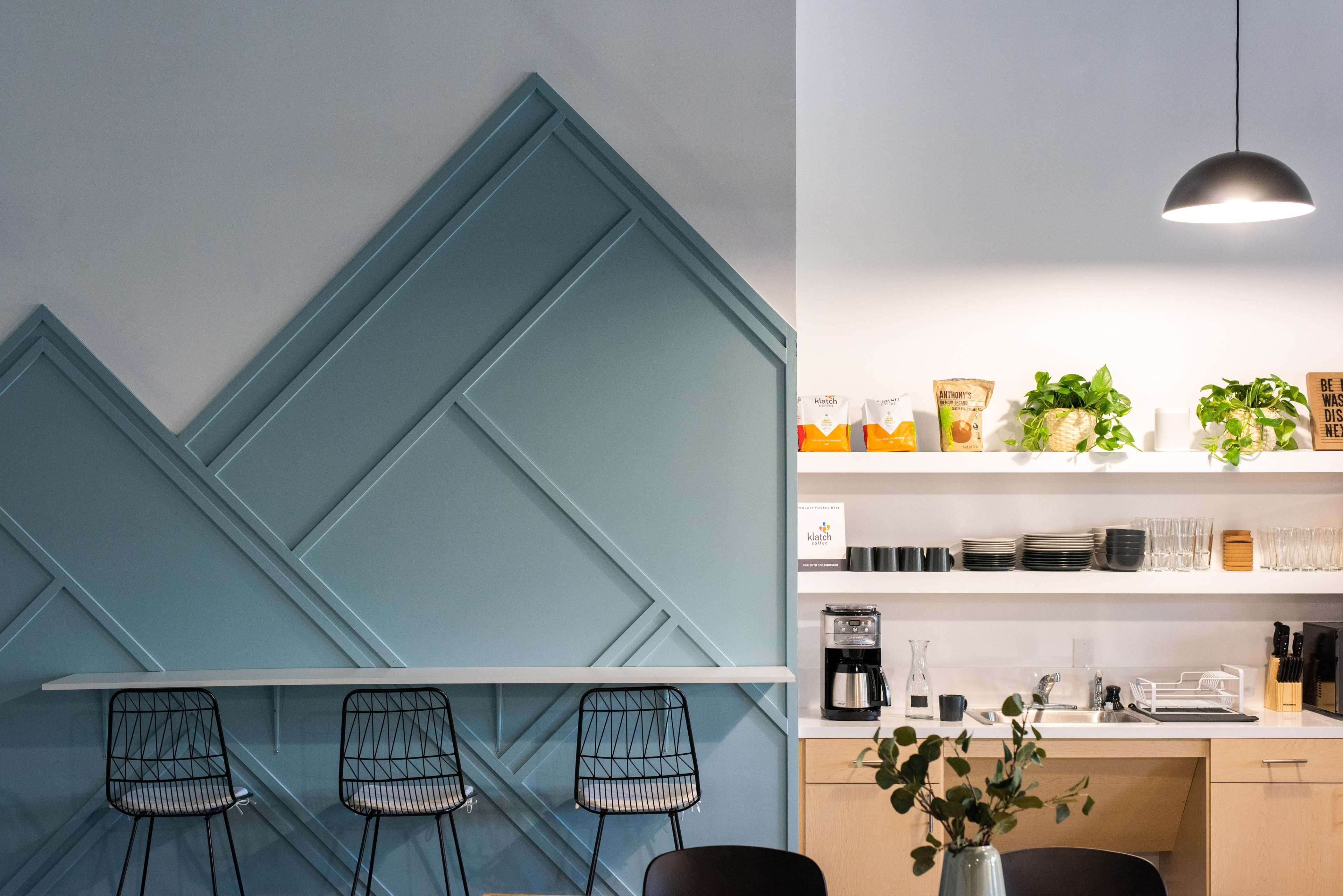 A modern café interior features a geometric blue wall, a white counter with black wire stools, and a shelf displaying various plants and kitchen items.