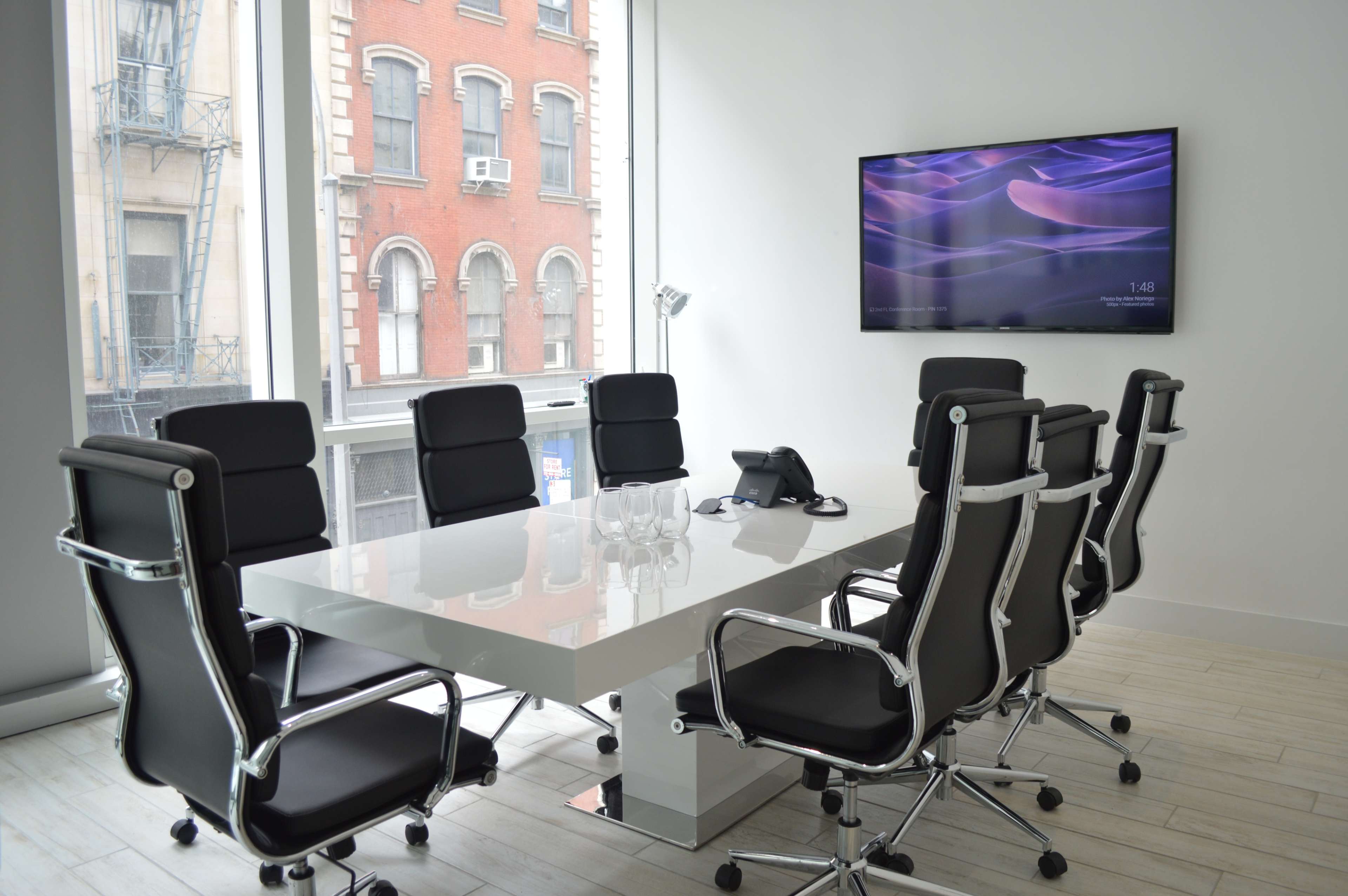 A modern conference room features a large white table surrounded by black ergonomic chairs, with a wall-mounted screen and large windows showing an urban view outside.